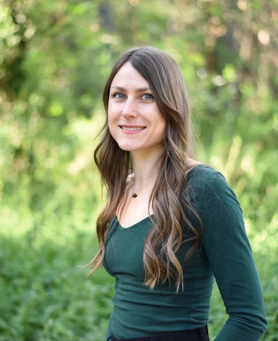A woman with long wavy brown hair and blue eyes standing outdoors with green foliage in the background.