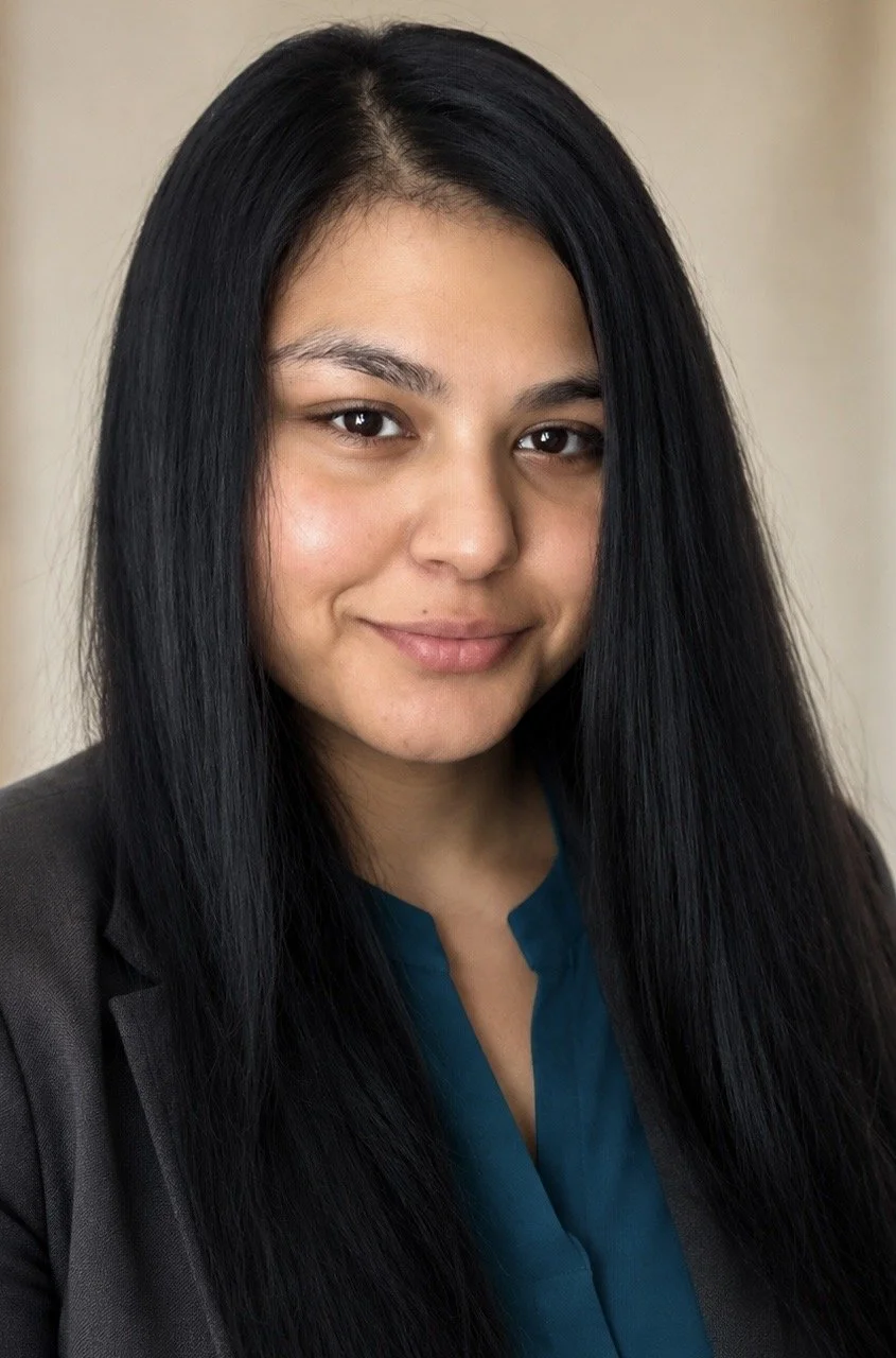 A woman with long black hair, wearing a dark blazer over a teal blouse, smiling softly at the camera.