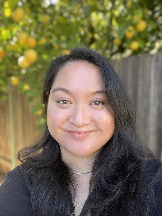 A woman with long dark hair smiling outdoors with lemon trees and a wooden fence in the background.