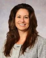 Portrait of a woman with long brown hair, smiling, wearing a gray blazer.
