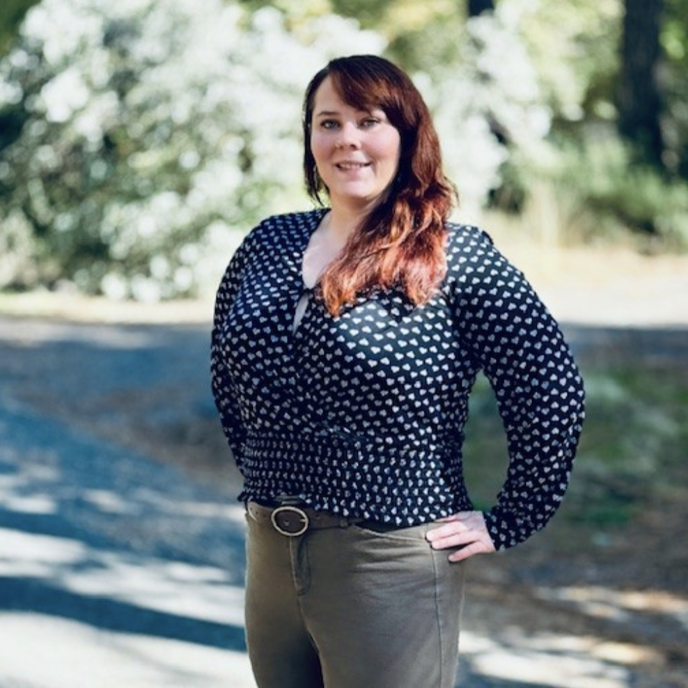 A woman standing outdoors on a sunny day with trees in the background. She has long, reddish-brown hair and is wearing a navy blouse with small white heart patterns, tan pants, and a belt. She is smiling and has one hand on her hip.