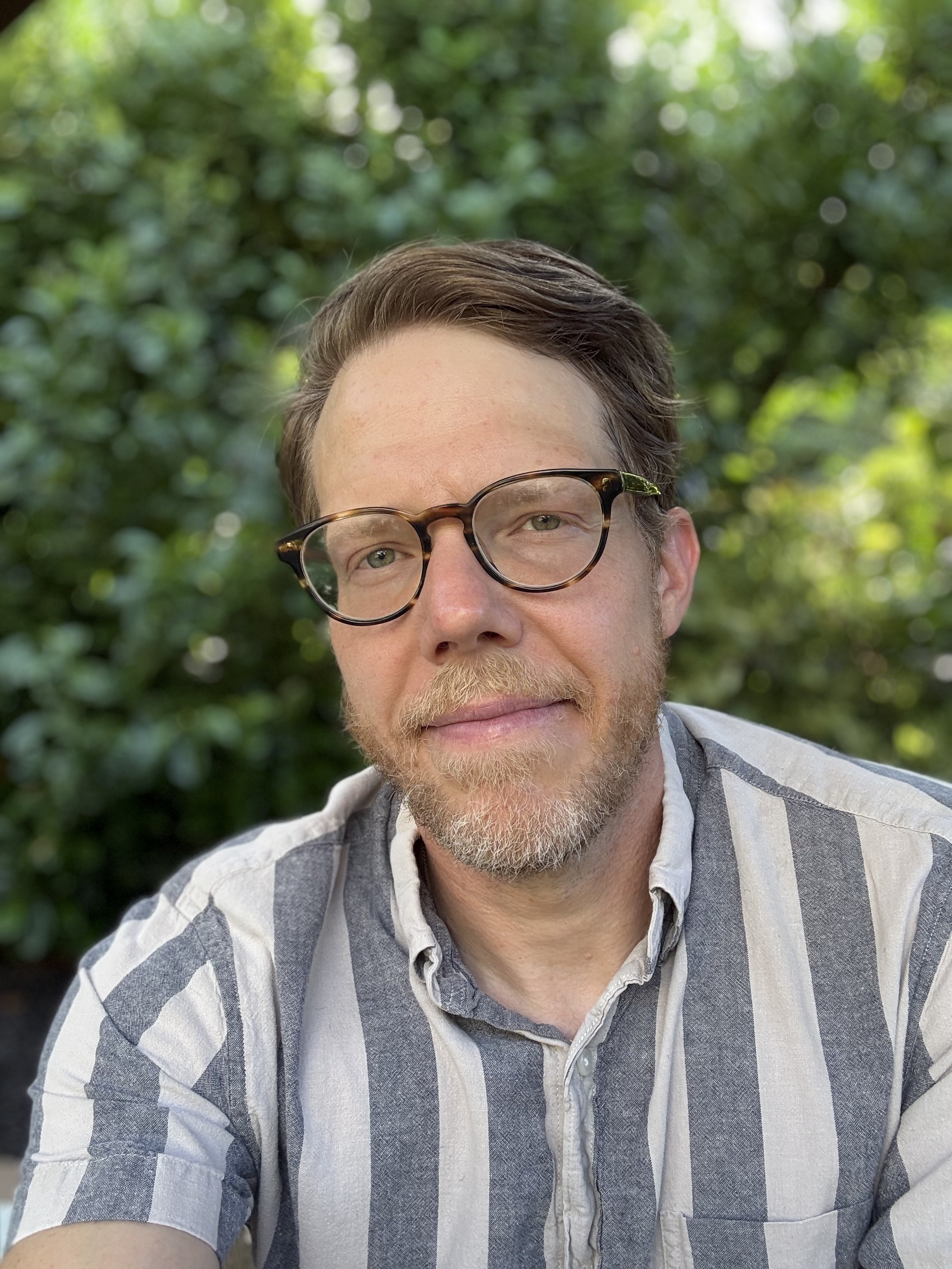 A man with glasses and a beard, wearing a striped shirt, outdoors in front of a blurred, green, leafy background.