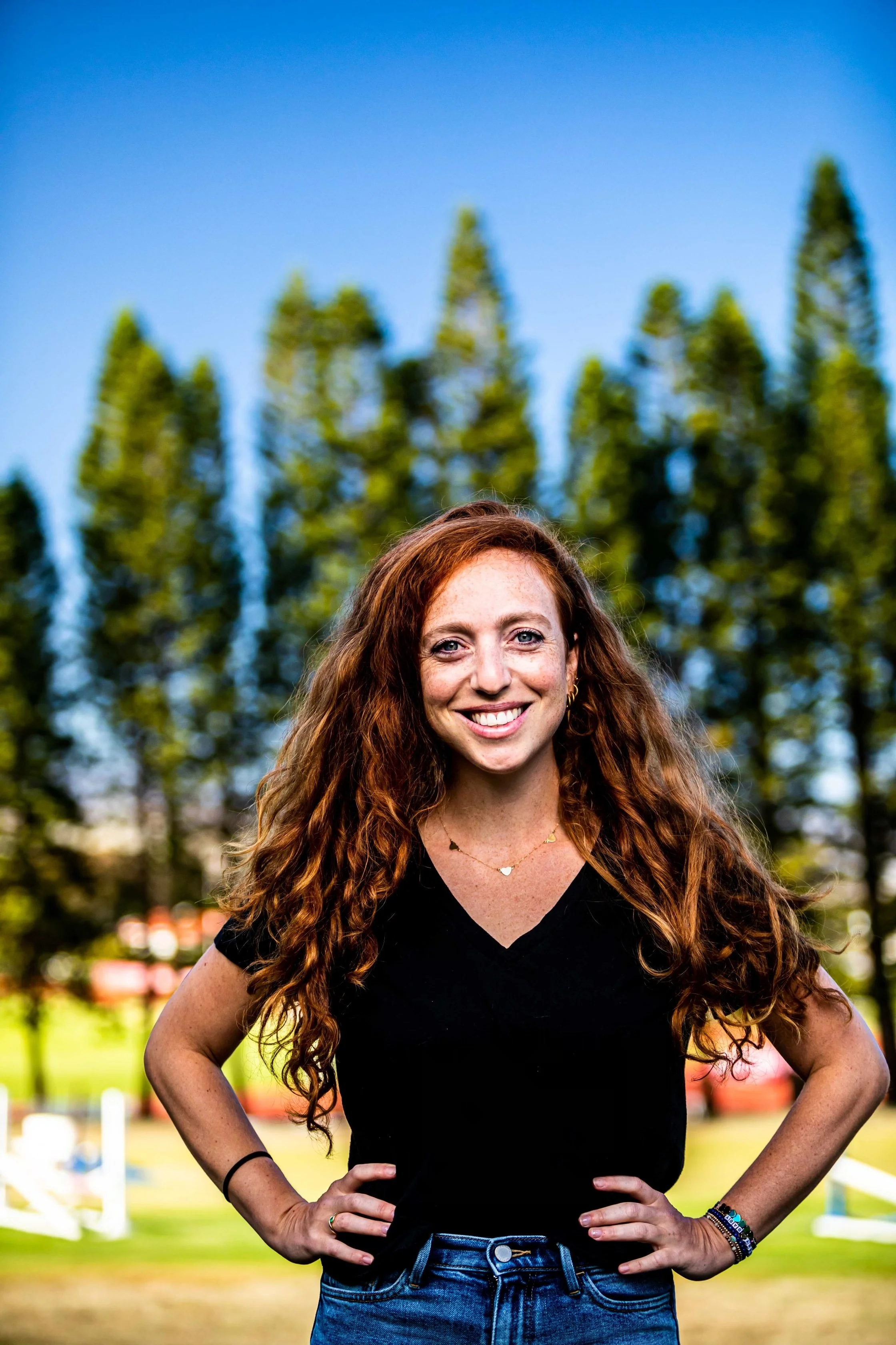 A woman with long curly red hair smiling outdoors, wearing a black V-neck top, blue jeans, and jewelry, with trees and a clear blue sky in the background.