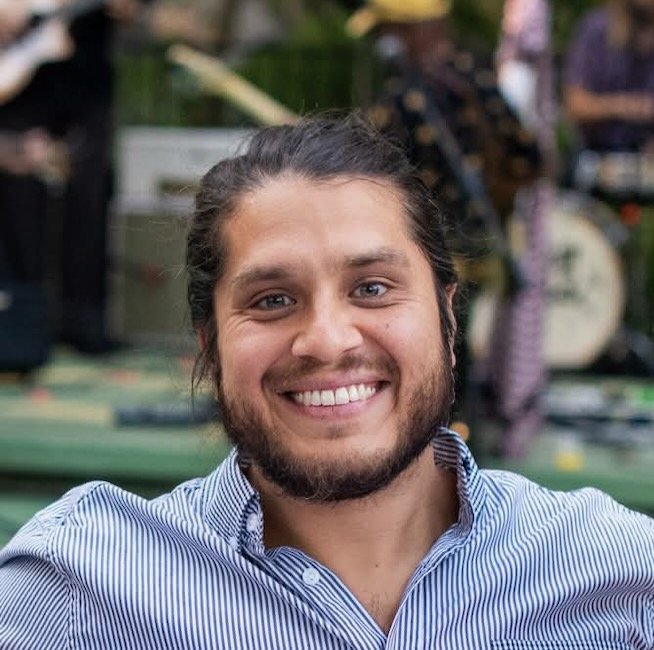 Smiling man with long hair and beard wearing a striped shirt, outdoor setting with blurred background.