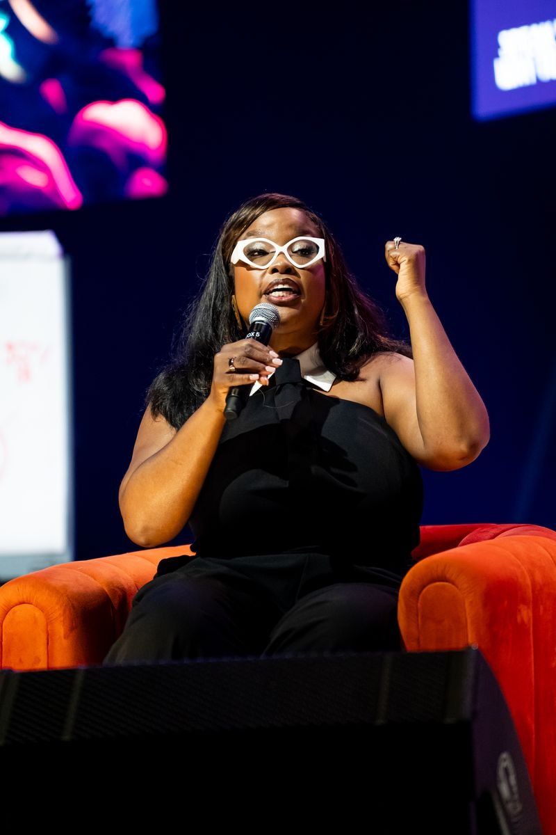 A woman with long dark hair, wearing white glasses and a black outfit, is sitting on an orange chair on stage, holding a microphone and gesturing with a raised fist.