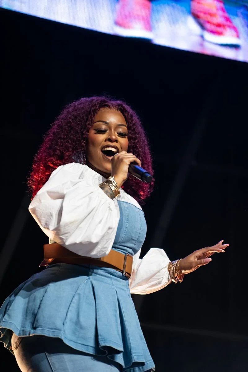 A woman singing into a microphone on stage, wearing a blue dress with a tan belt, white puffed sleeves, and jewelry, with a large screen in the background.