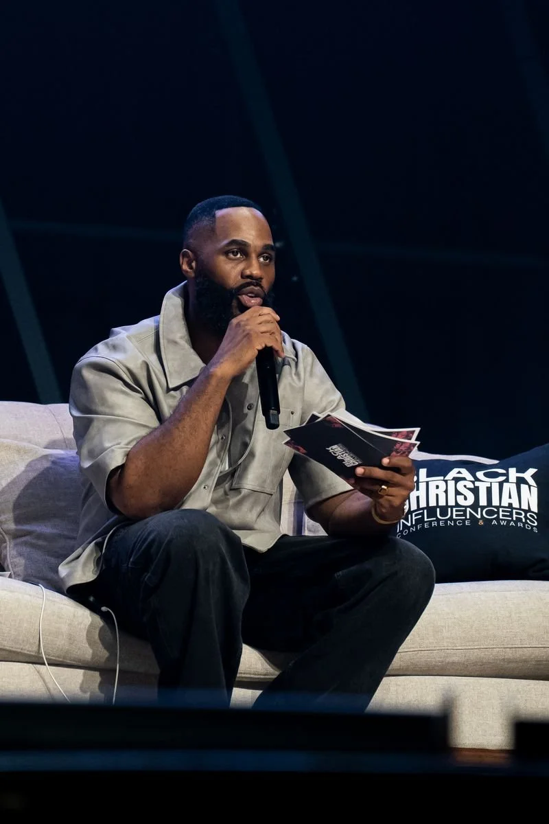 A man with a beard and short hair sitting on a beige sofa, holding a microphone in one hand and a booklet in the other, speaking at a Christian conference or event.