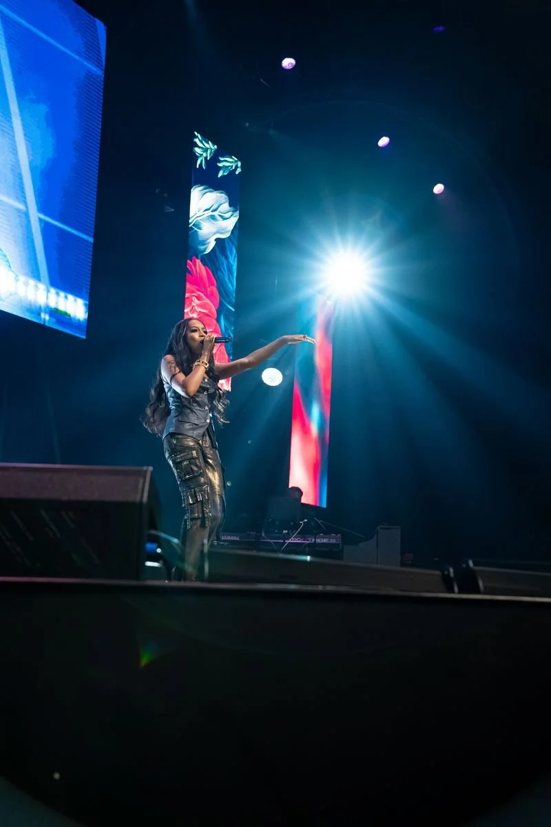 A woman performing on stage with stage lights and digital screens behind her, singing into a microphone.