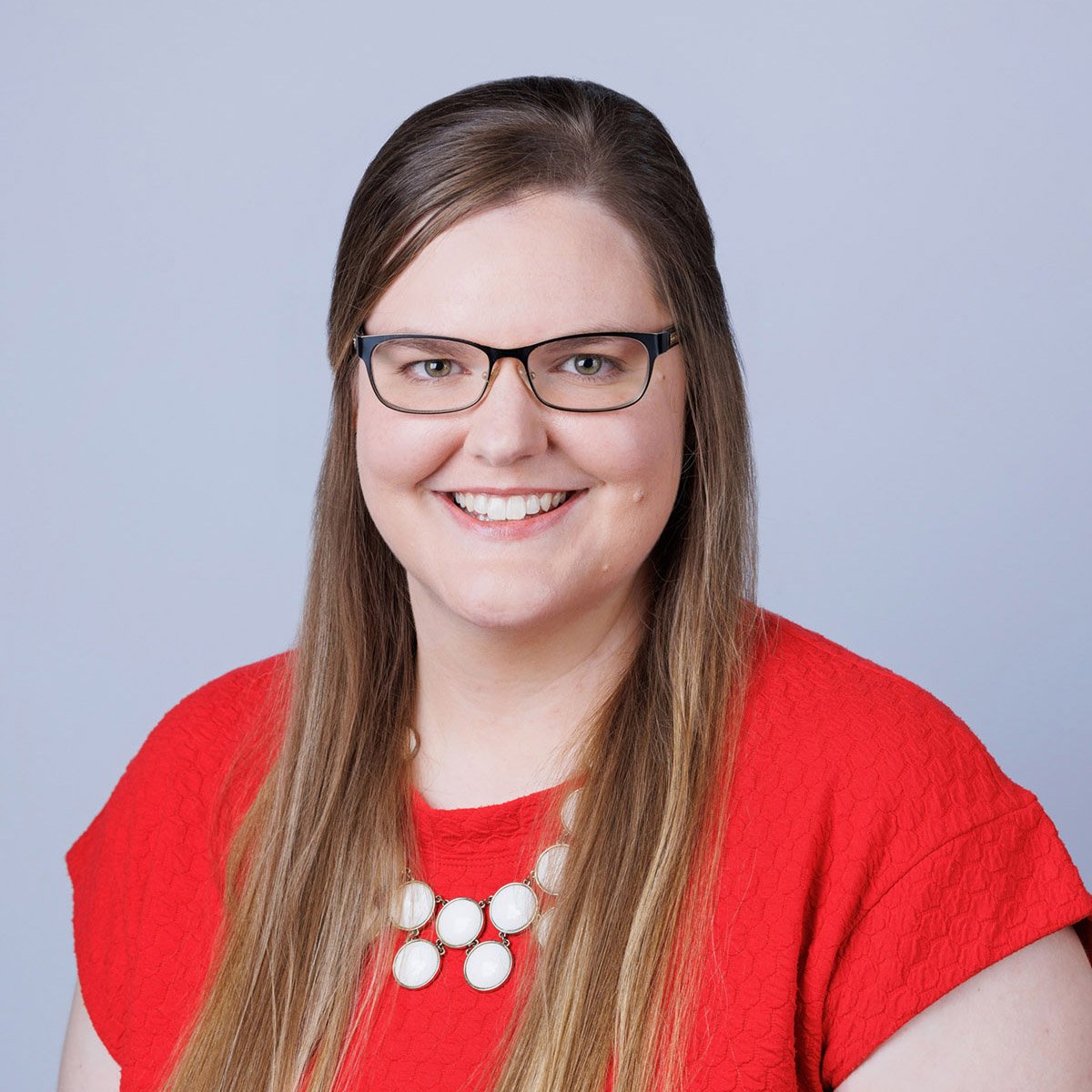 A woman with long brown hair wearing glasses, a red top, and a white necklace, smiling at the camera against a light grey background.