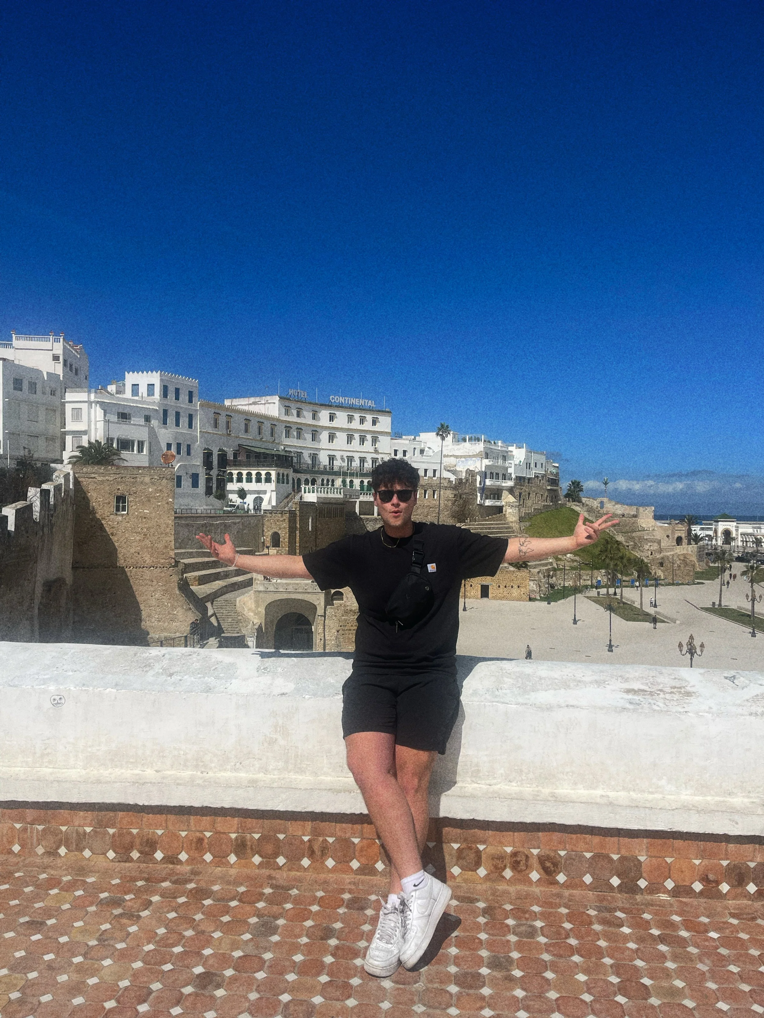 A young man in black clothing and sunglasses posing with arms outstretched in front of white buildings and a sandy area under a bright blue sky.