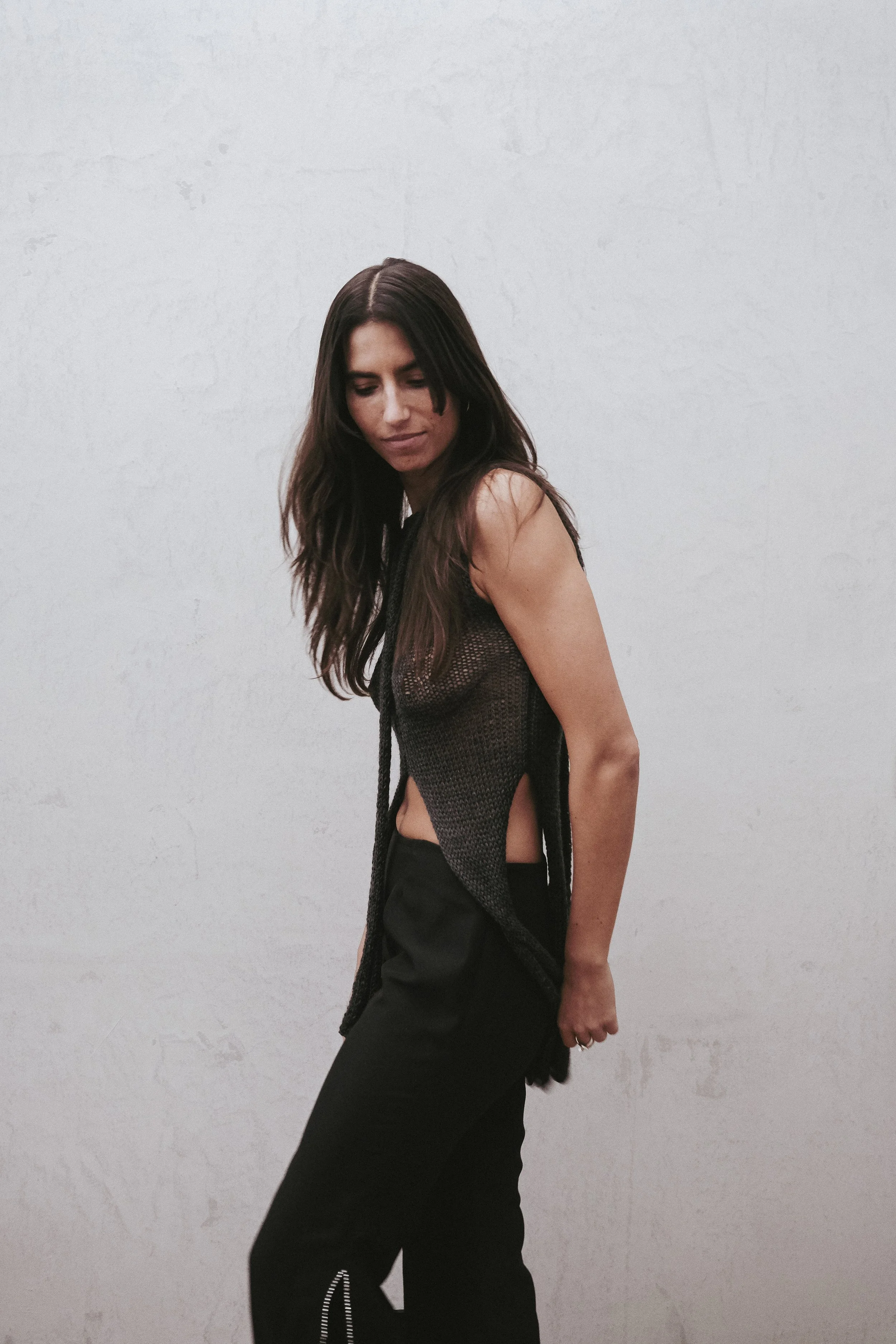 A woman with long dark hair wearing a black sleeveless top and black pants standing against a plain light-colored wall, looking down with a relaxed pose.