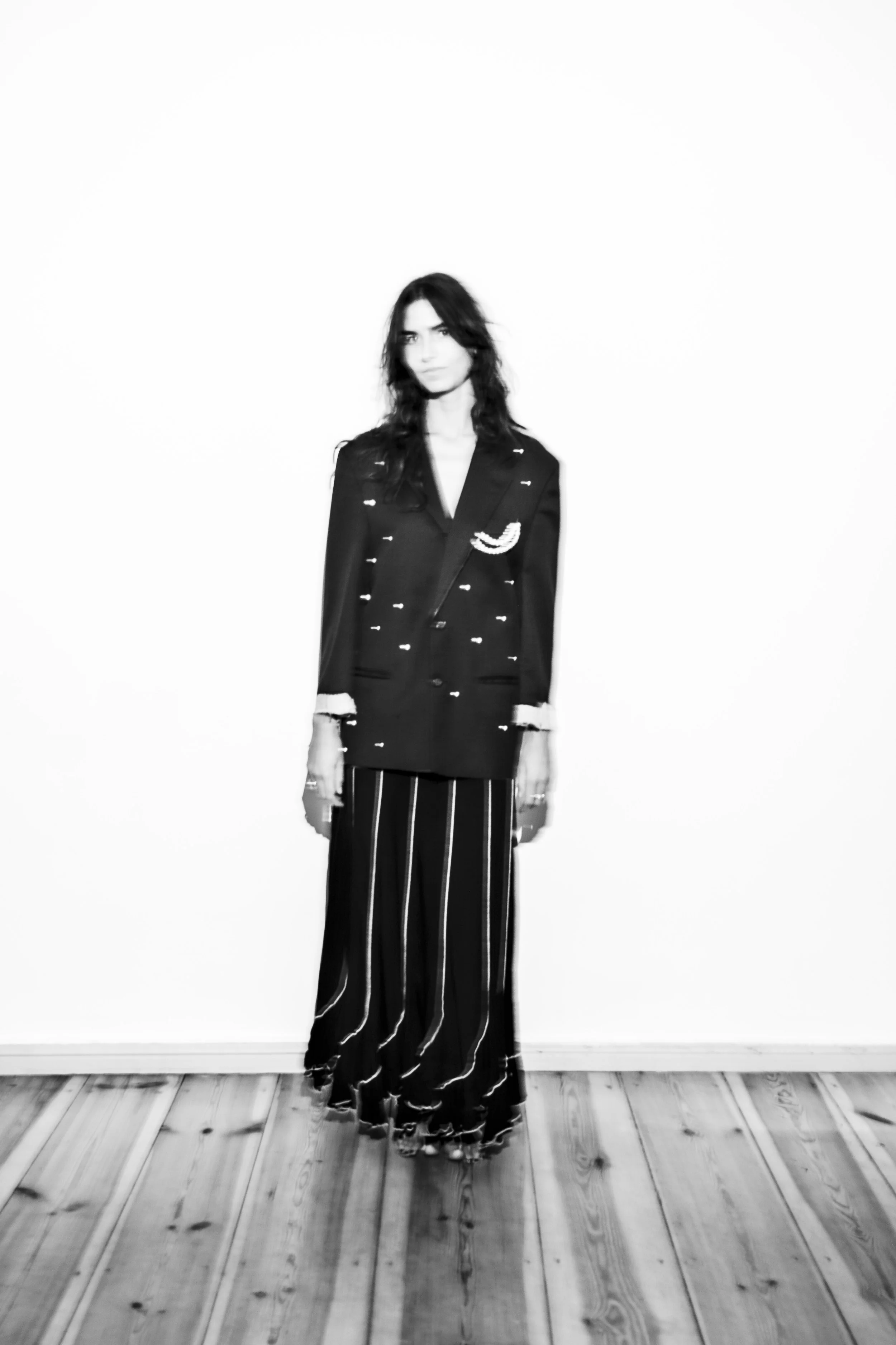 A woman standing against a plain white background, wearing a dark blazer with light-colored embellishments and wide-leg striped pants, with long wavy hair and a serious expression.