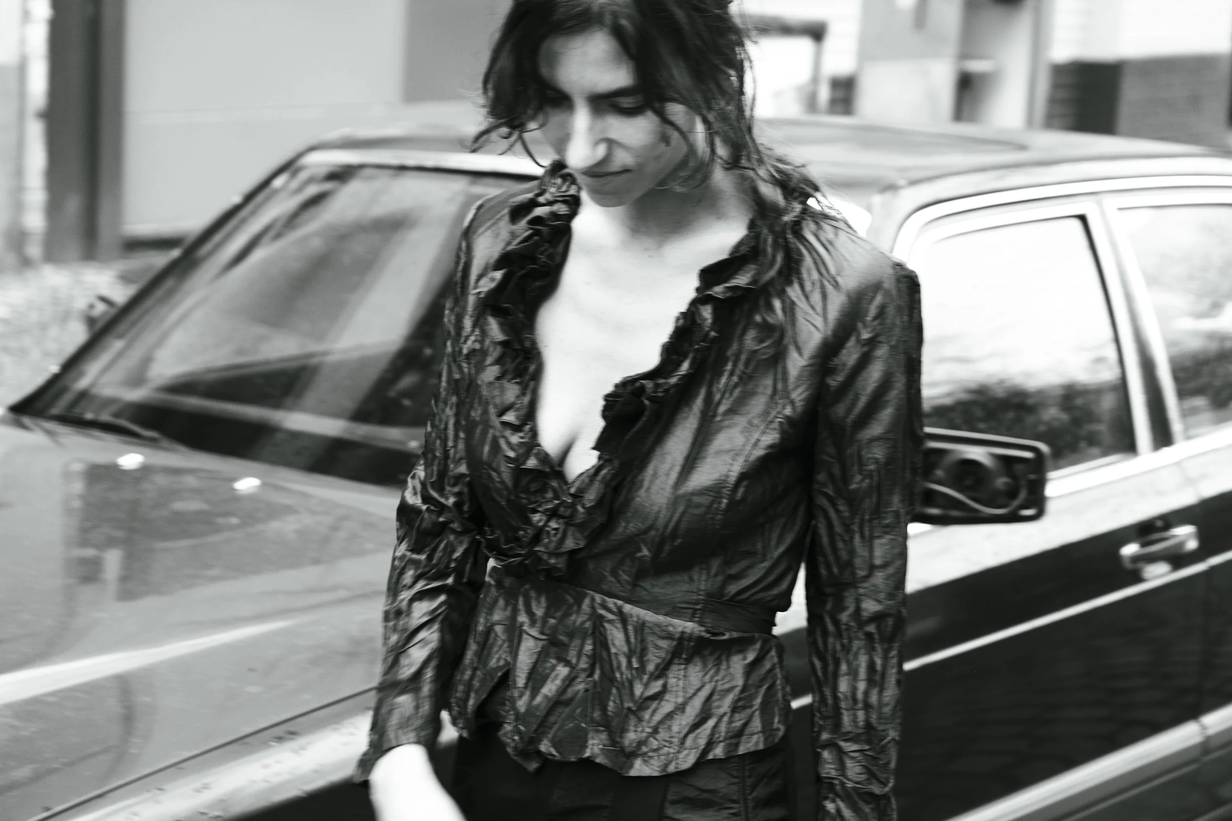 A woman with dark, wavy hair stands in front of a car on a city street, wearing a shiny black leather jacket and black pants.