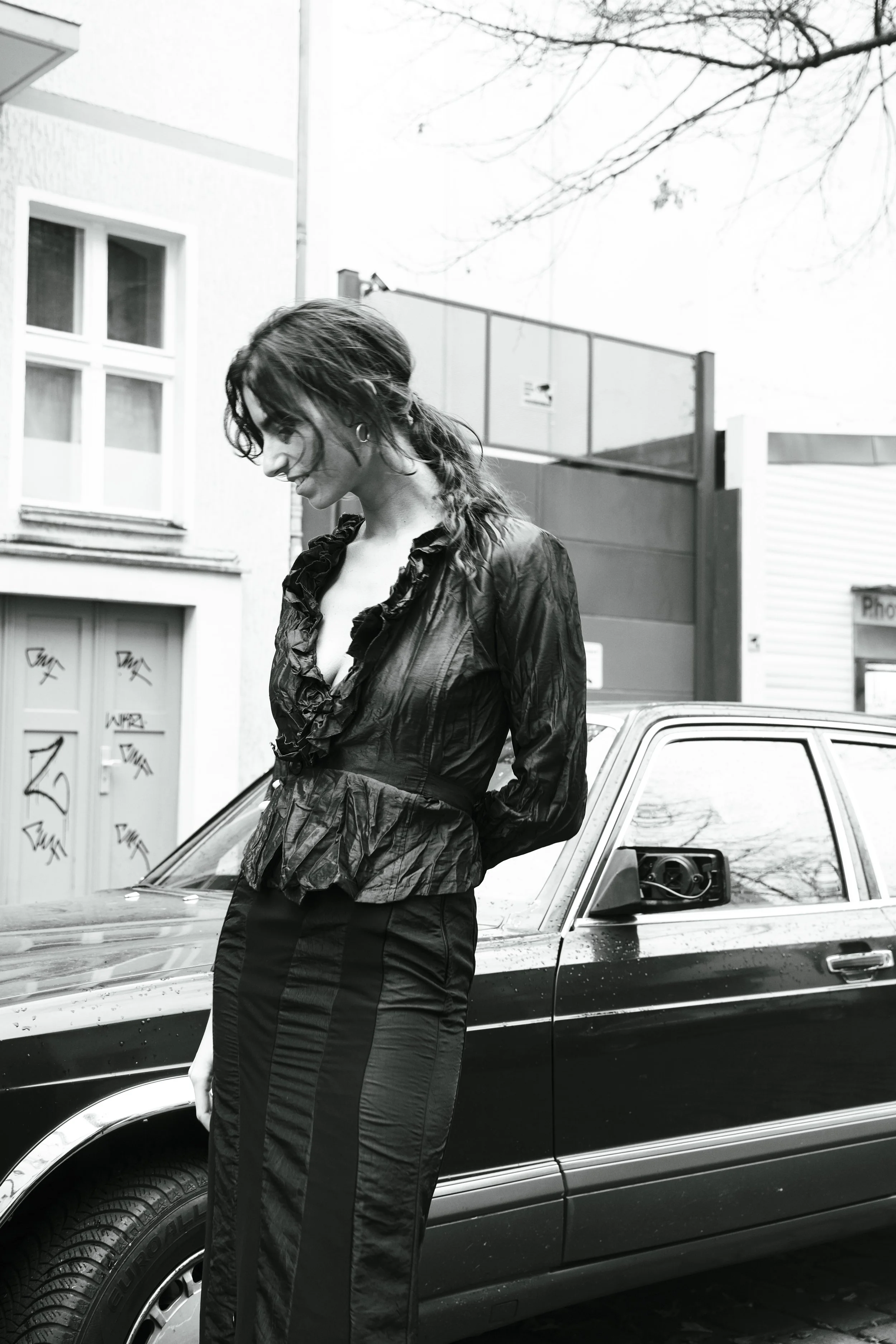 A woman with wavy hair wearing a ruffled, shiny jacket and striped pants, standing outdoors next to a black car. The background shows a building with graffiti on the door and some trees.