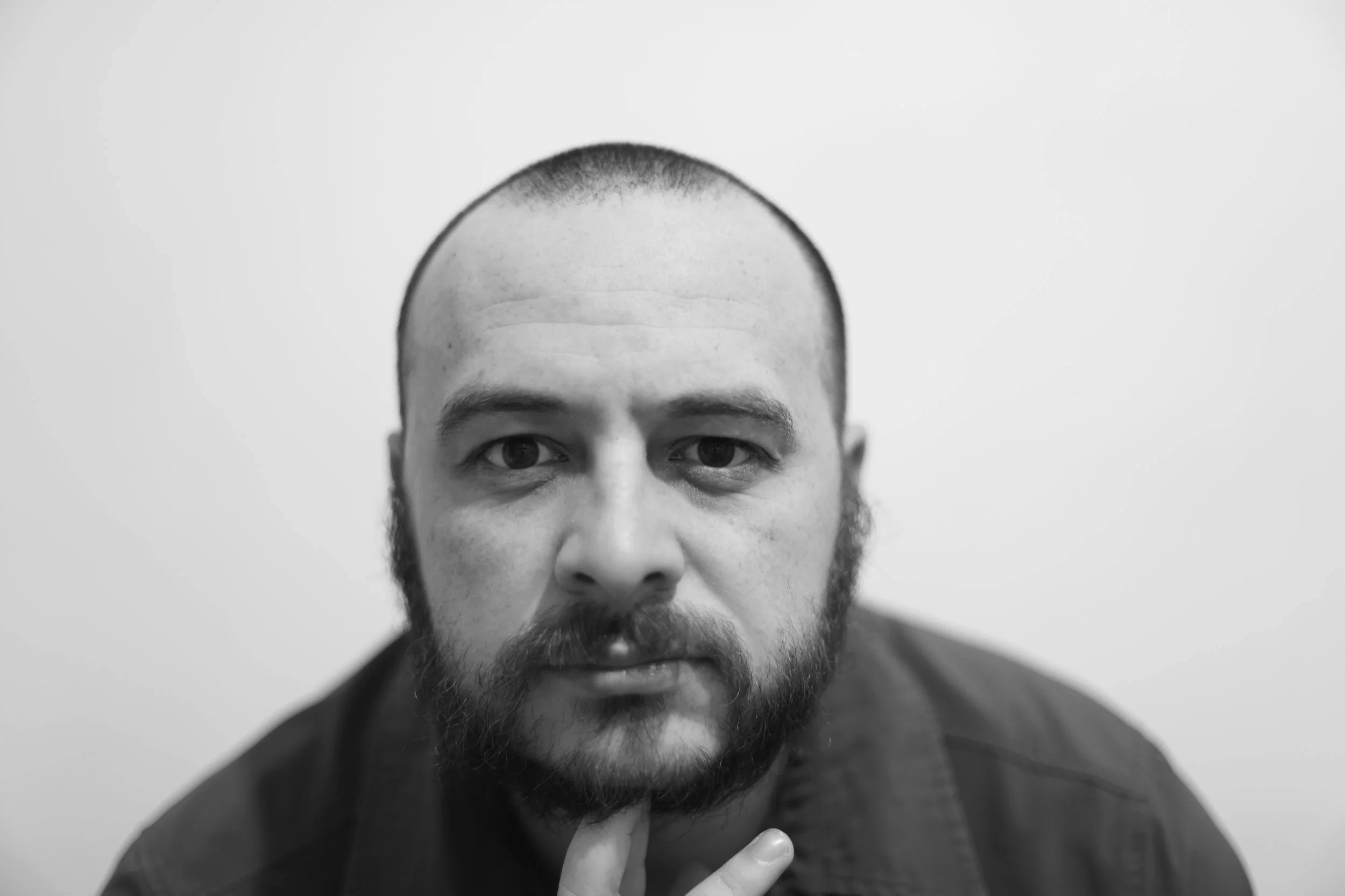 A black and white close-up portrait of a man with a beard and short hair, looking directly at the camera with a thoughtful expression, resting his chin on his hand.