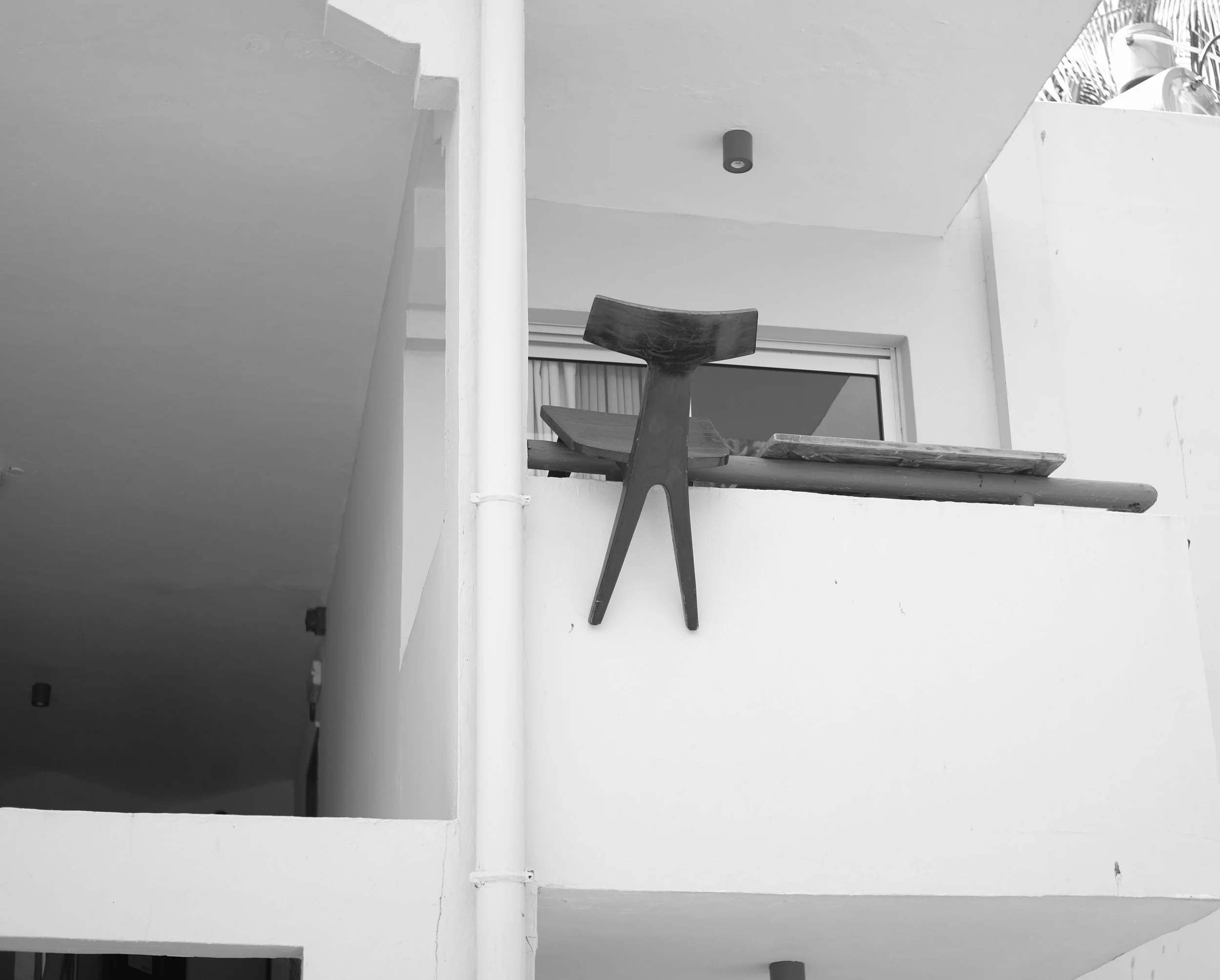 Black and white photo of a small wooden chair placed on the edge of a balcony or ledge, overlooking an interior space with a sliding glass door.