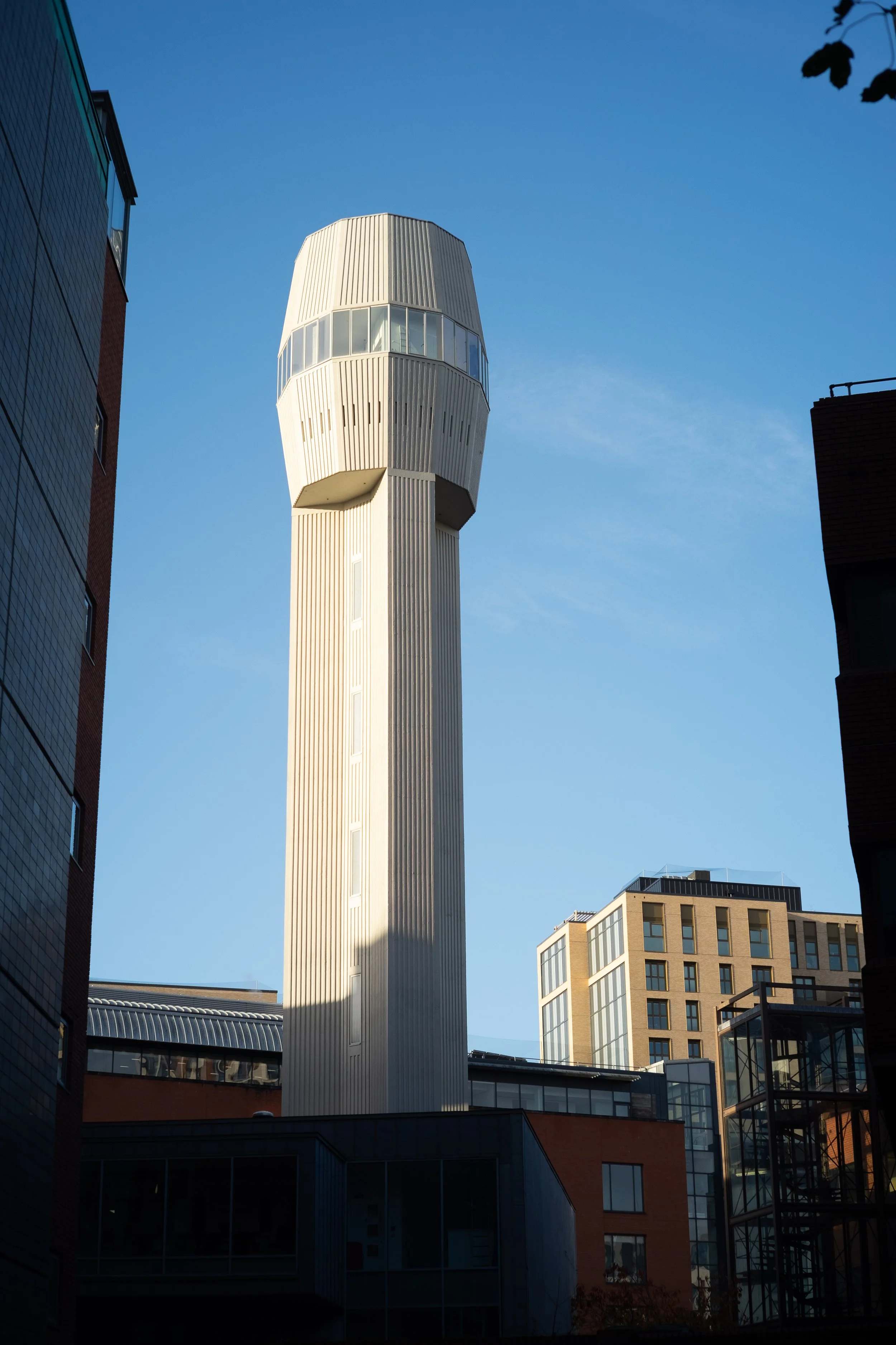 A tall, white, modern control tower or observation tower set against a clear blue sky, surrounded by buildings with glass windows.