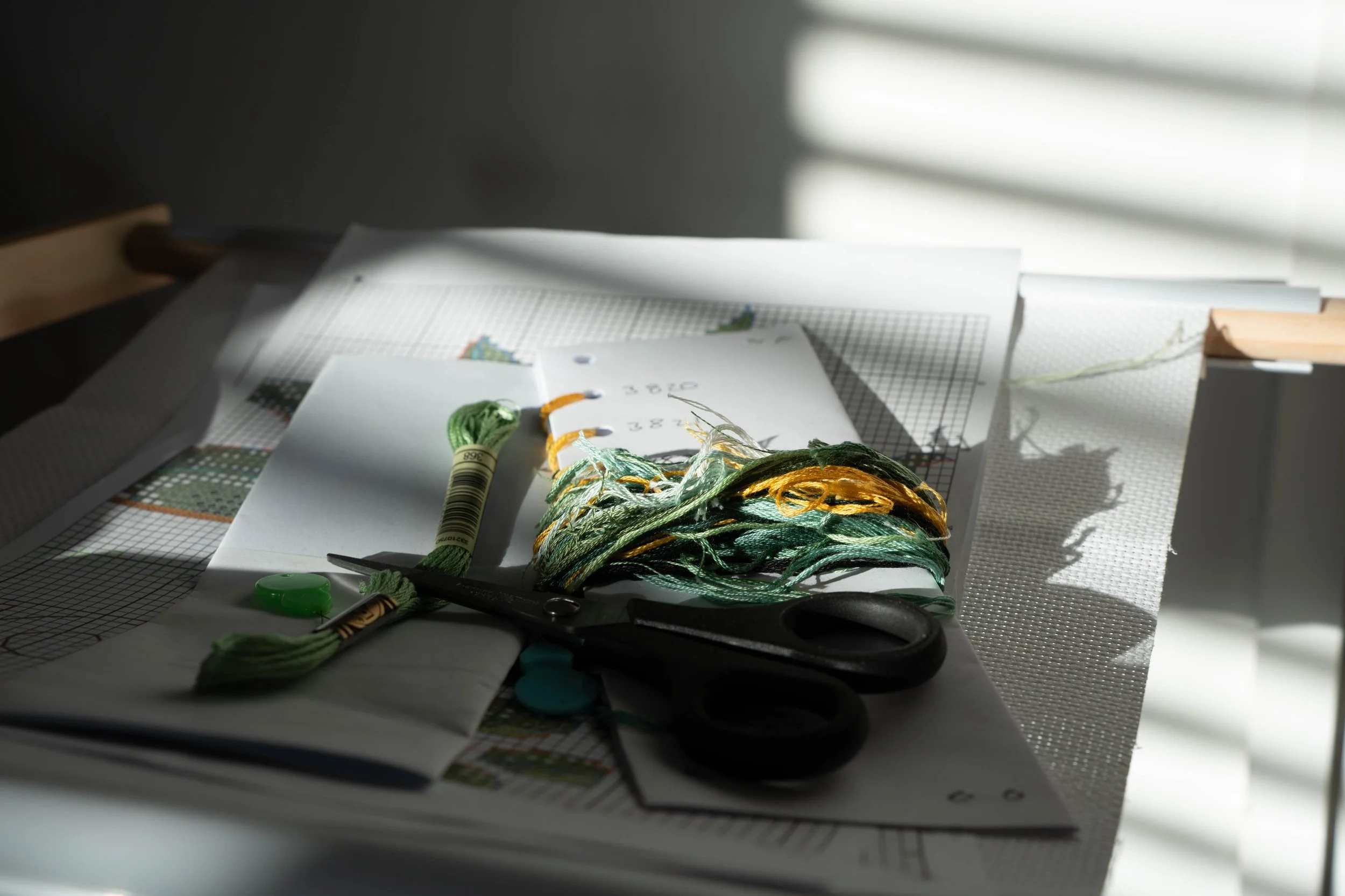 A crafting workspace with embroidery floss, scissors, and fabric on a table, illuminated by partial sunlight and cast shadows.