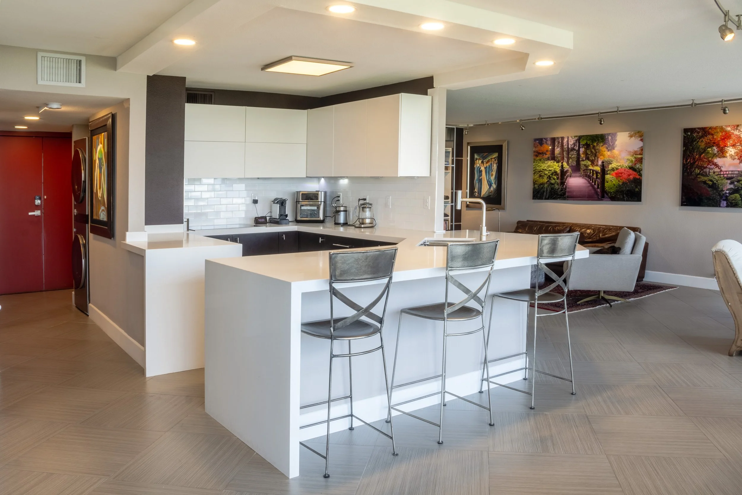 Modern open-concept kitchen with white cabinetry, stainless steel appliances, and a large white island with three metal barstools. Adjacent living area features armchairs, a leather sofa, and landscape artwork on the walls.