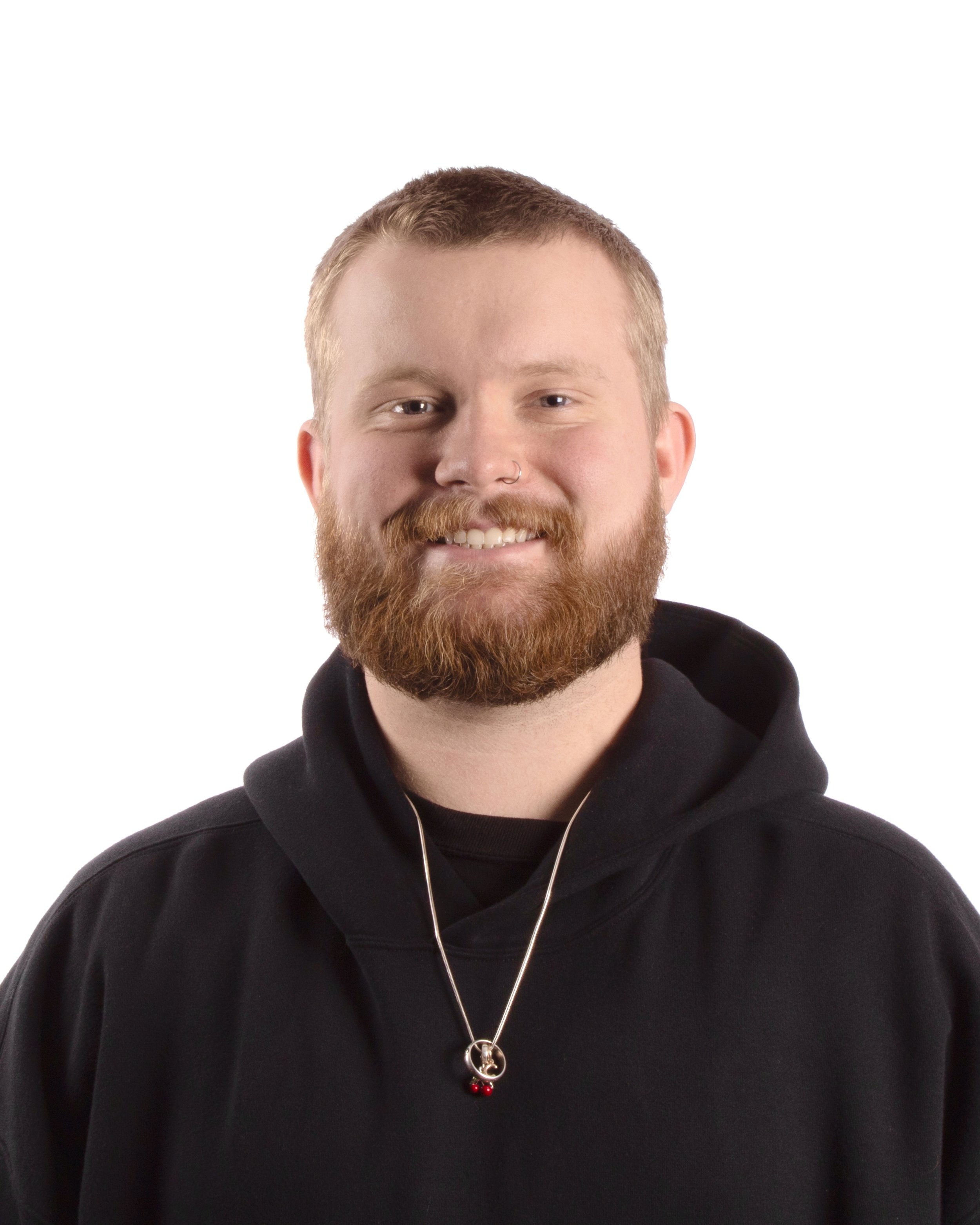 Portrait of a smiling man with a red beard, wearing a black hoodie and a necklace with a pendant, against a white background.
