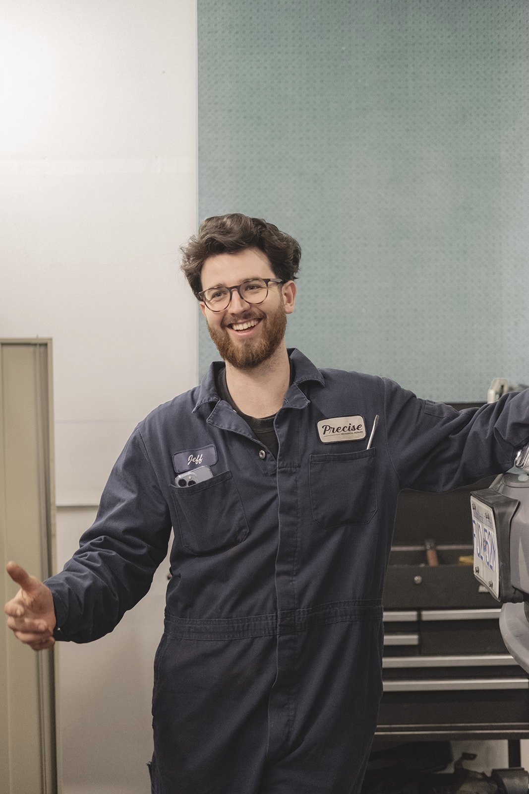 A smiling man with glasses and a beard wearing a navy blue work uniform with a name tag that reads 'Jeff' and a patch that says 'Precise', standing next to a vending machine in a workshop or repair shop.