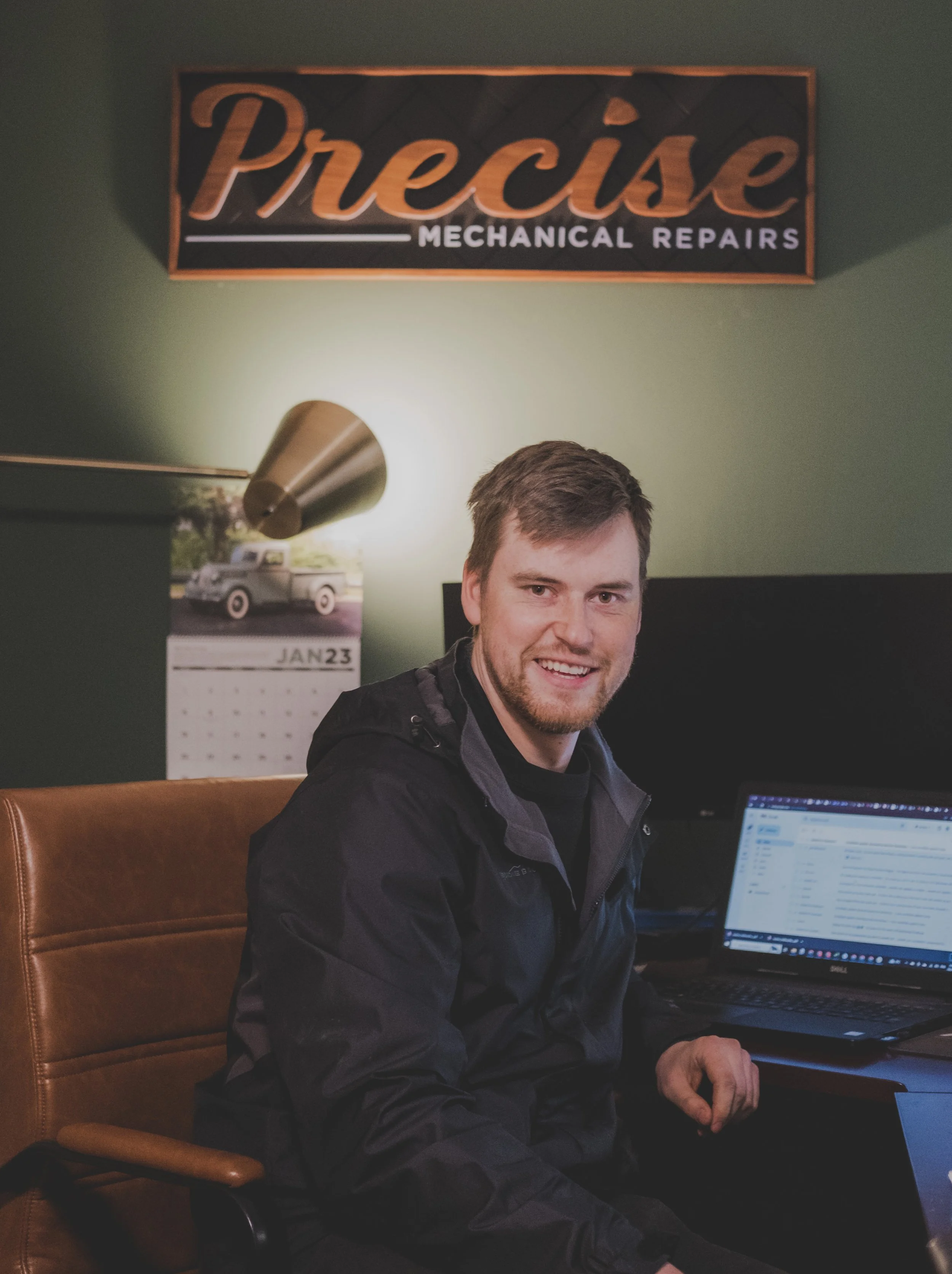 A smiling man sitting at a desk with a laptop in a room with a sign that says 'Precise Mechanical Repairs' on a green wall.
