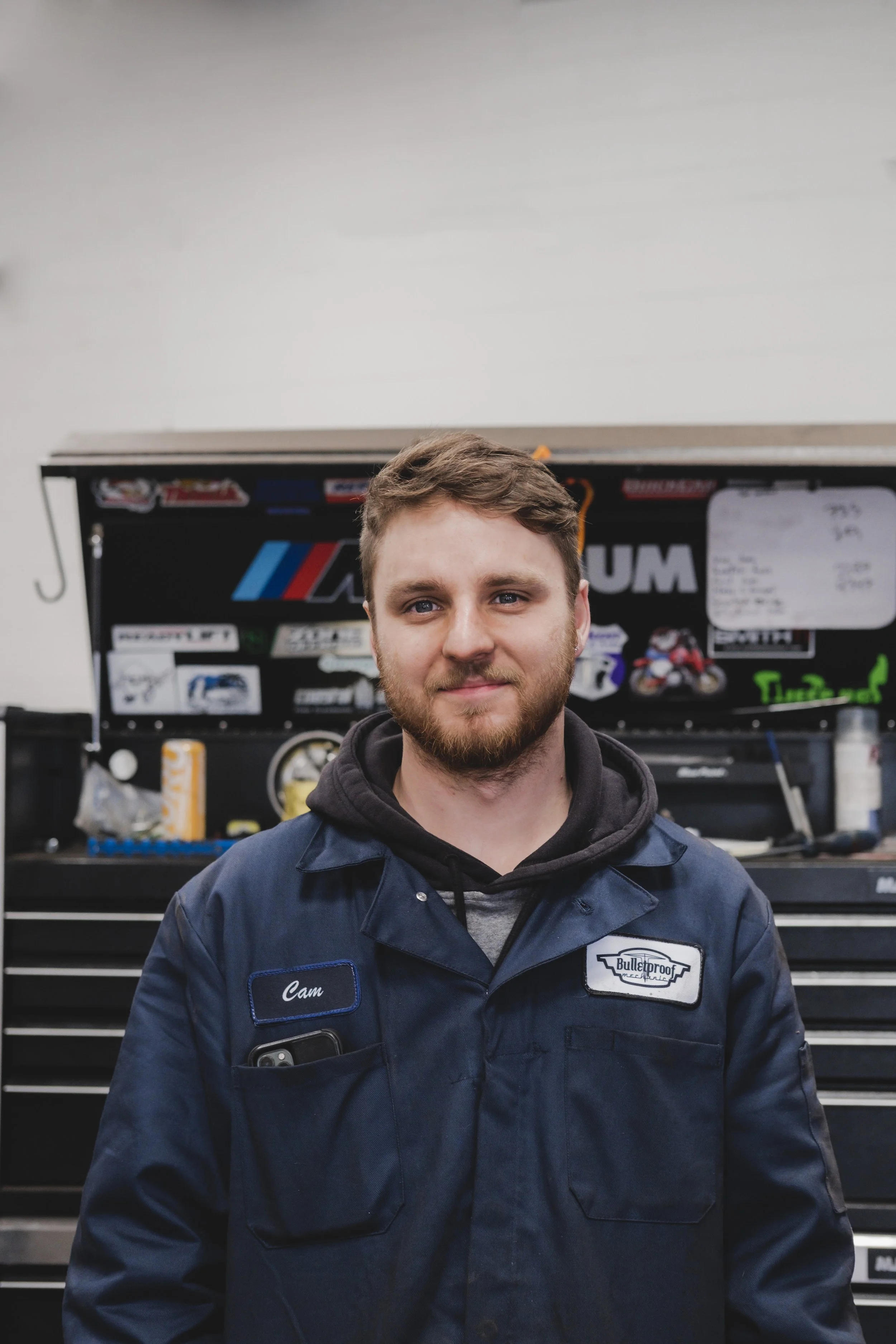 A young man with a beard, wearing a dark blue work jacket with a name tag that says 'Cam' and a patch that says 'Bulletproof', standing in front of a tool chest with stickers and a whiteboard.