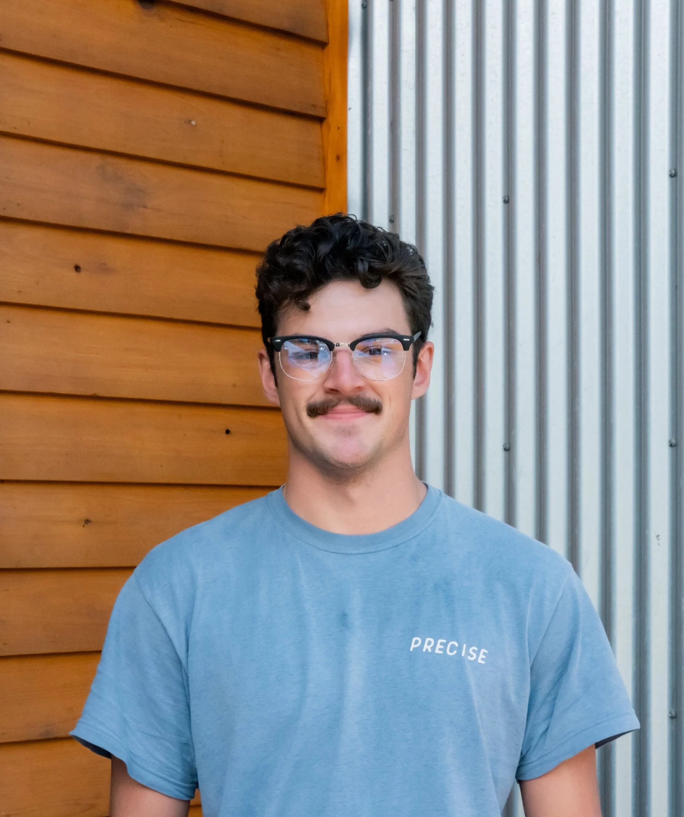 A young man with dark curly hair, glasses, and a mustache standing in front of a wooden and metal wall, wearing a blue t-shirt with the word 'PRECISE' printed on it.