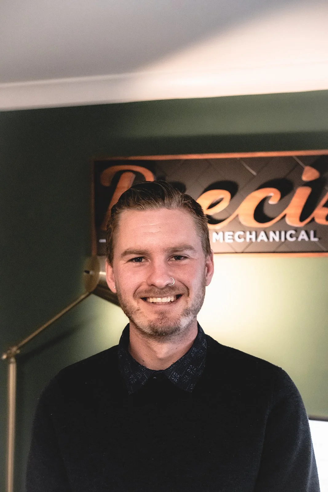 A young man with light brown hair, a nose ring, and a beard, smiling in front of a sign that reads 'Reclat Mechanical' inside a room with dark green walls.