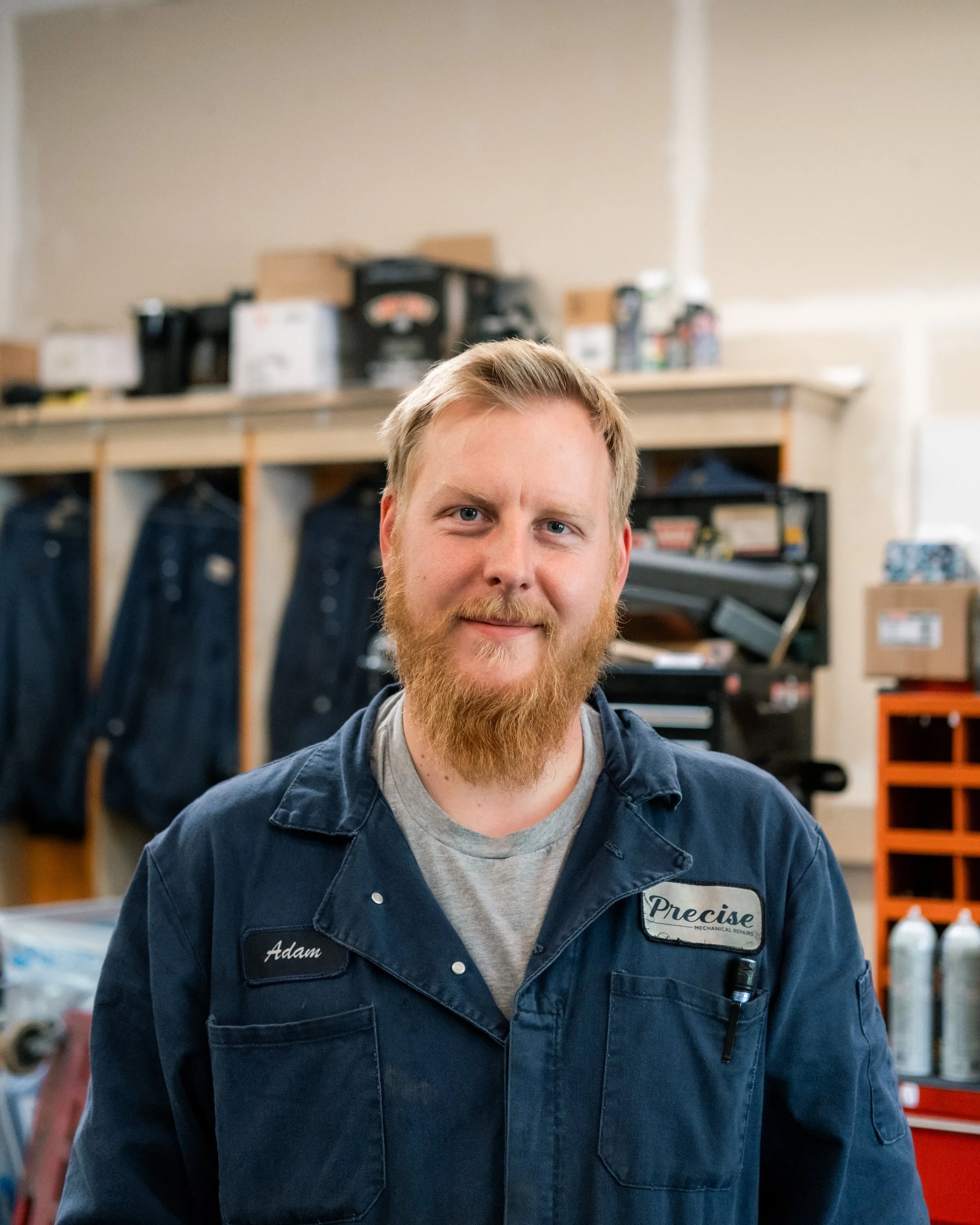A man with a beard and short blonde hair smiling in a workshop. He is wearing a navy blue work uniform with a name tag that reads "Adam" and another that says "Precise". Behind him, there are hooks with hanging dark blue jeans, shelves with tools, and storage boxes.