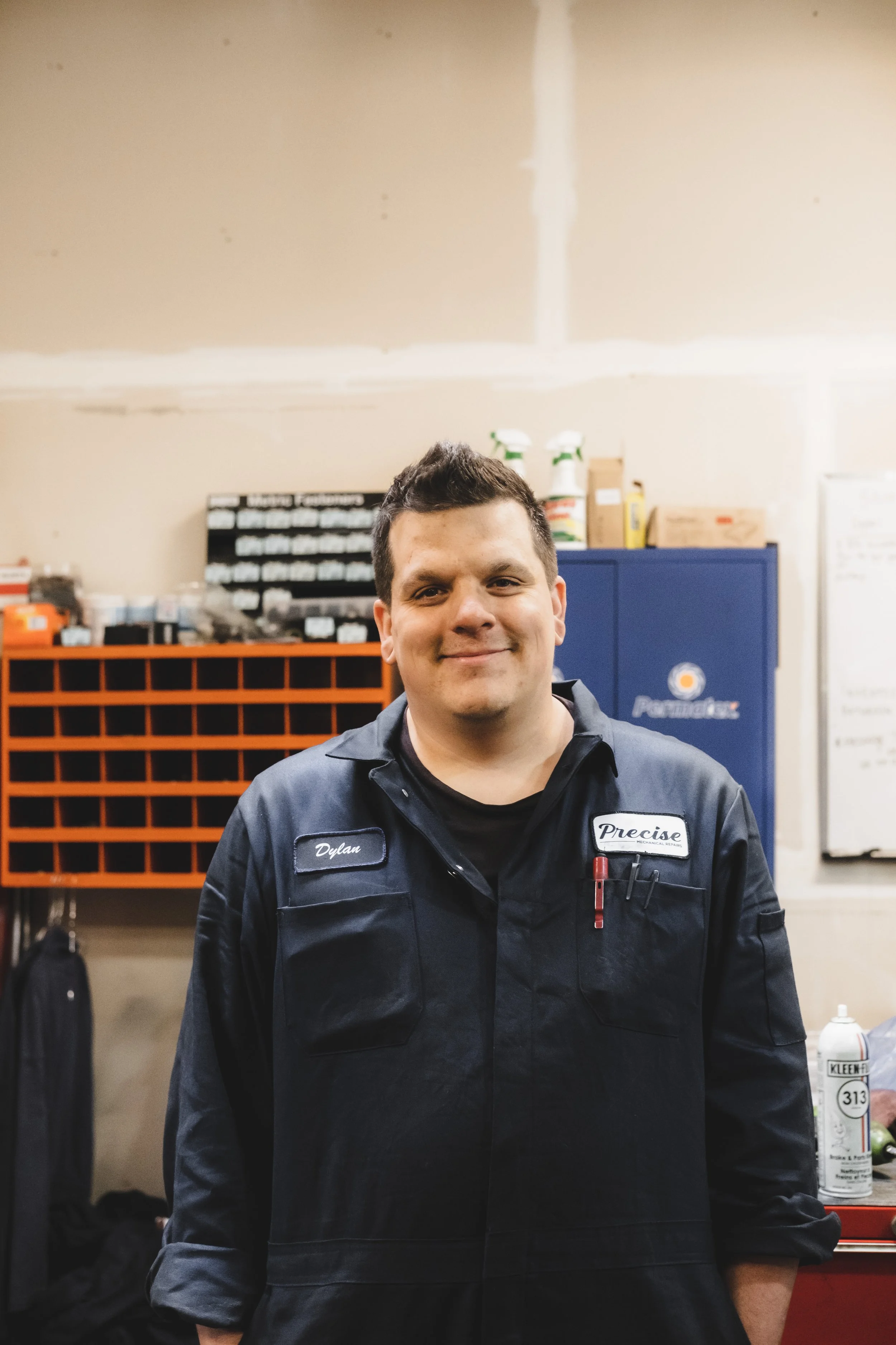 A smiling mechanic wearing a navy blue work uniform standing in a garage. The uniform has a name patch that reads 'Dylan' and a logo patch that says 'Precise.' In the background, there are organized shelves with tools, spray bottles, and a whiteboard on the wall.