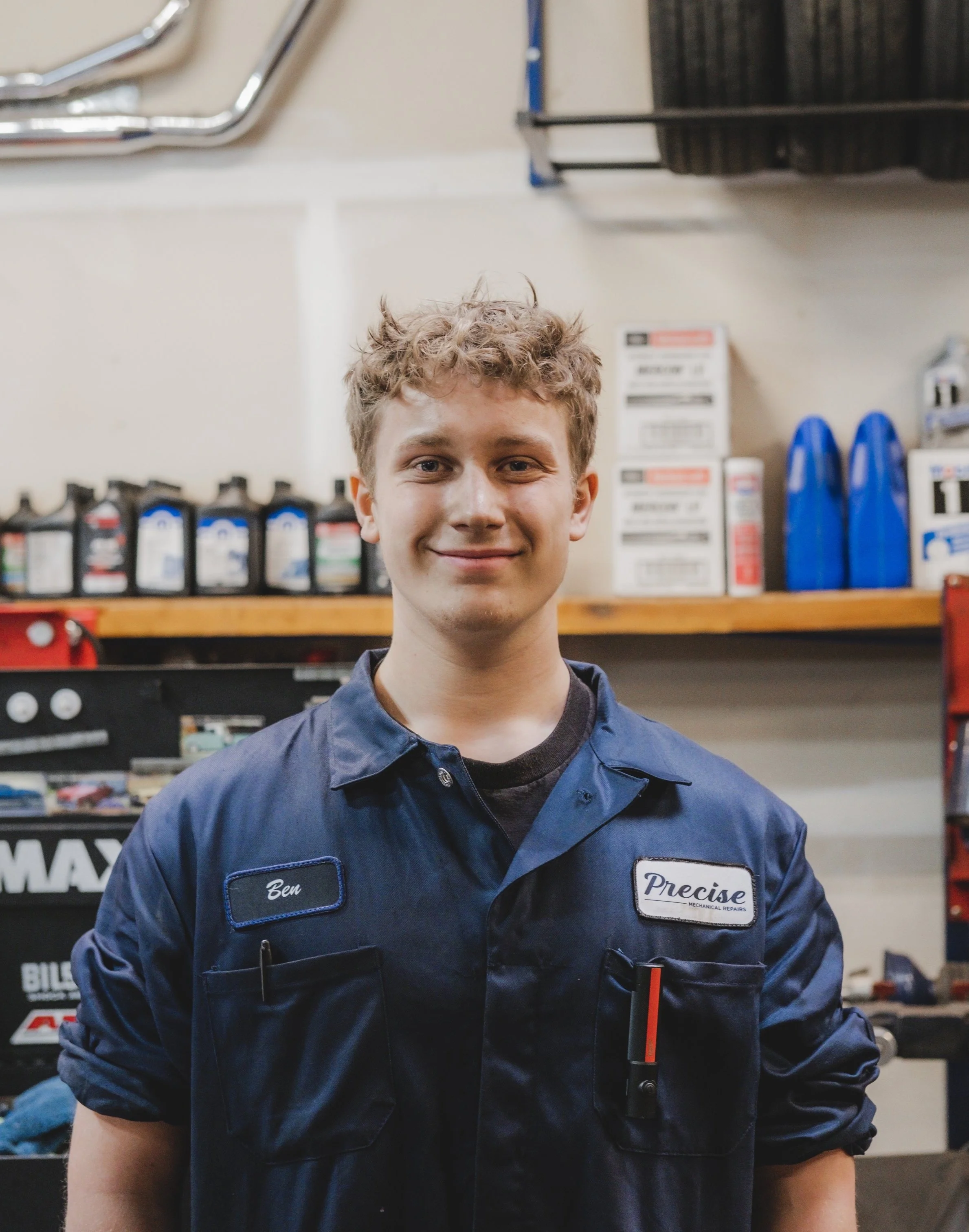A young man with curly hair smiling in a garage or auto repair shop, wearing a blue mechanic's uniform with name tag 'Ben' and a patch 'Precise'.
