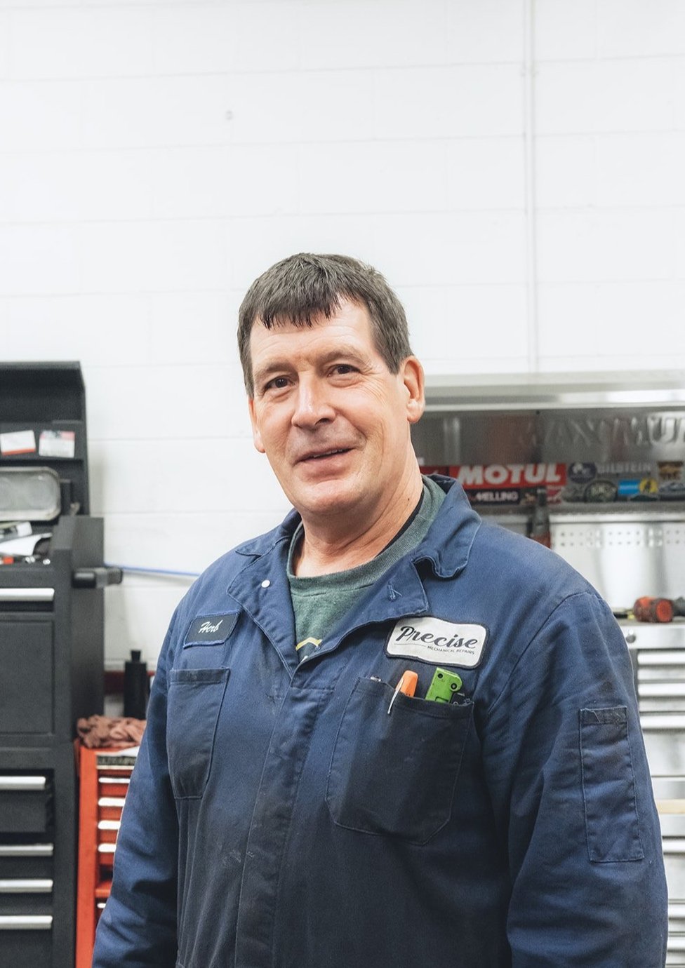 A man with short brown hair and a blue mechanic uniform standing in a workshop with tools and equipment.