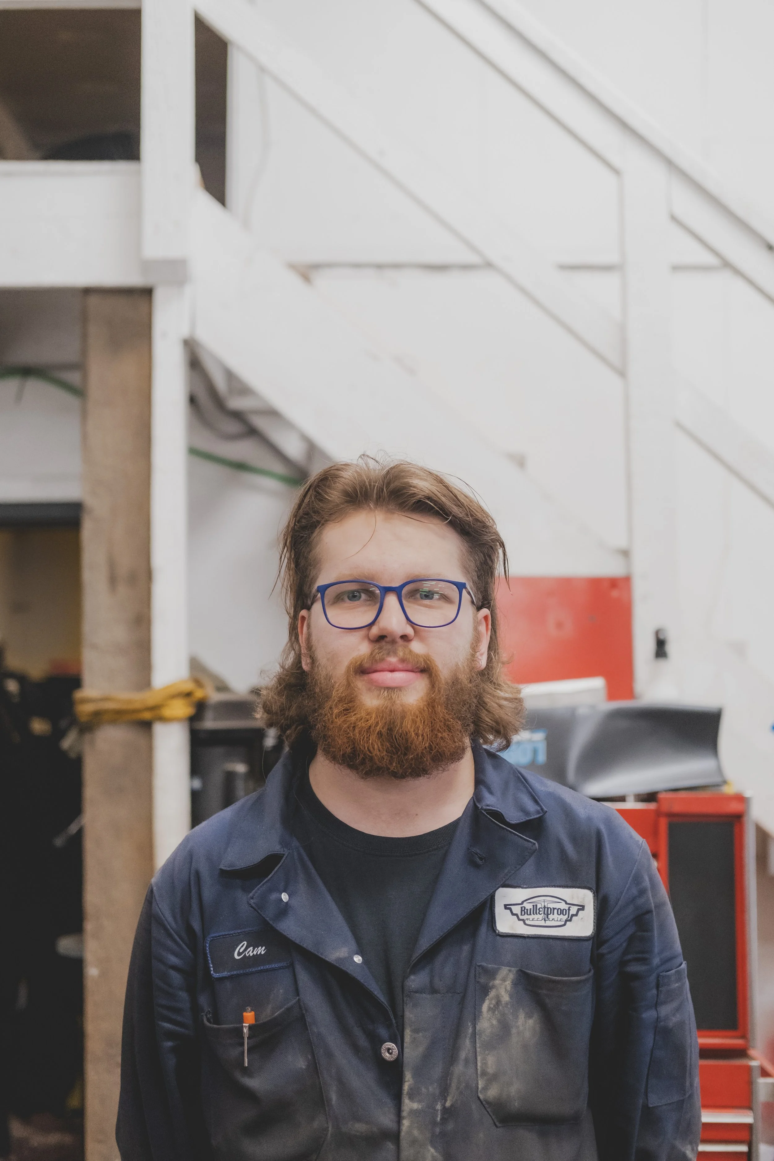A man with glasses and a beard wearing a blue work jumpsuit labeled 'Cam' and 'Bulletproof' standing in a workshop or garage.