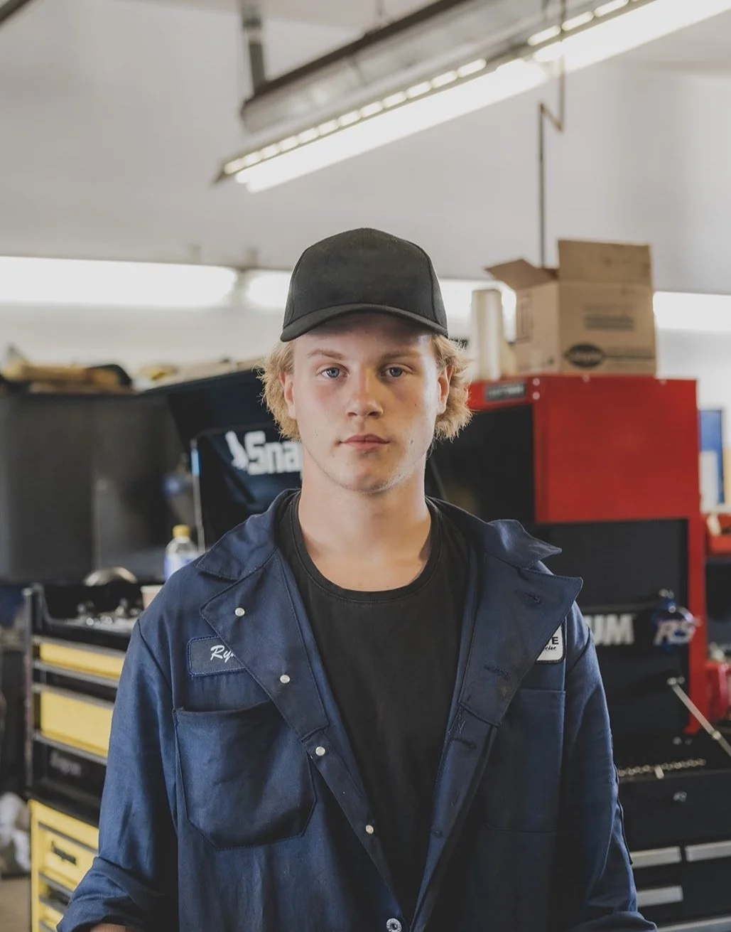 A young man with blonde hair wearing a black baseball cap, black t-shirt, and navy blue work jacket stands in a garage or workshop with toolboxes and cardboard boxes in the background.
