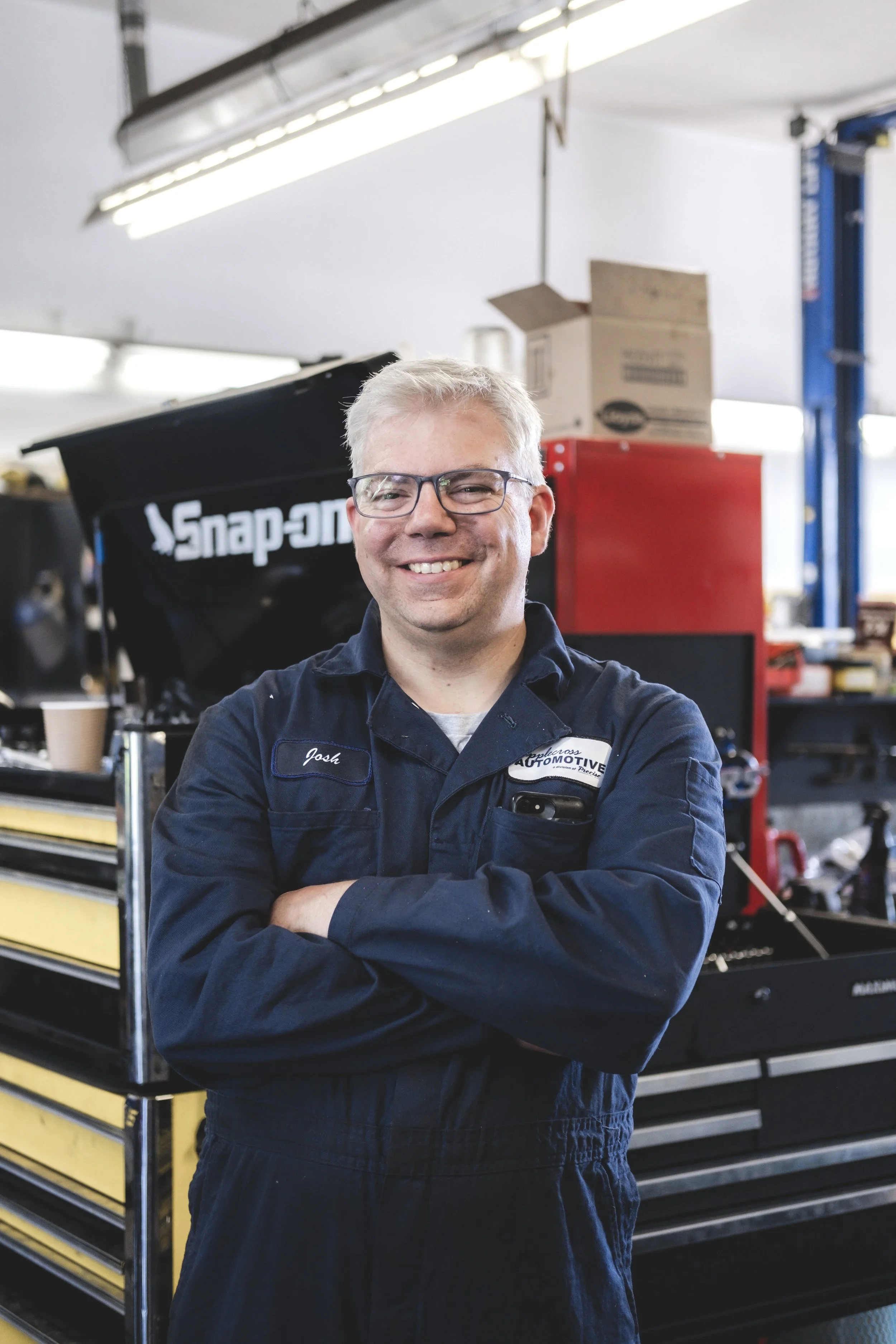A smiling man in a dark blue mechanic jumpsuit standing with arms crossed in an auto repair shop, surrounded by tools and equipment, including a Snap-on tool chest and red cabinets.
