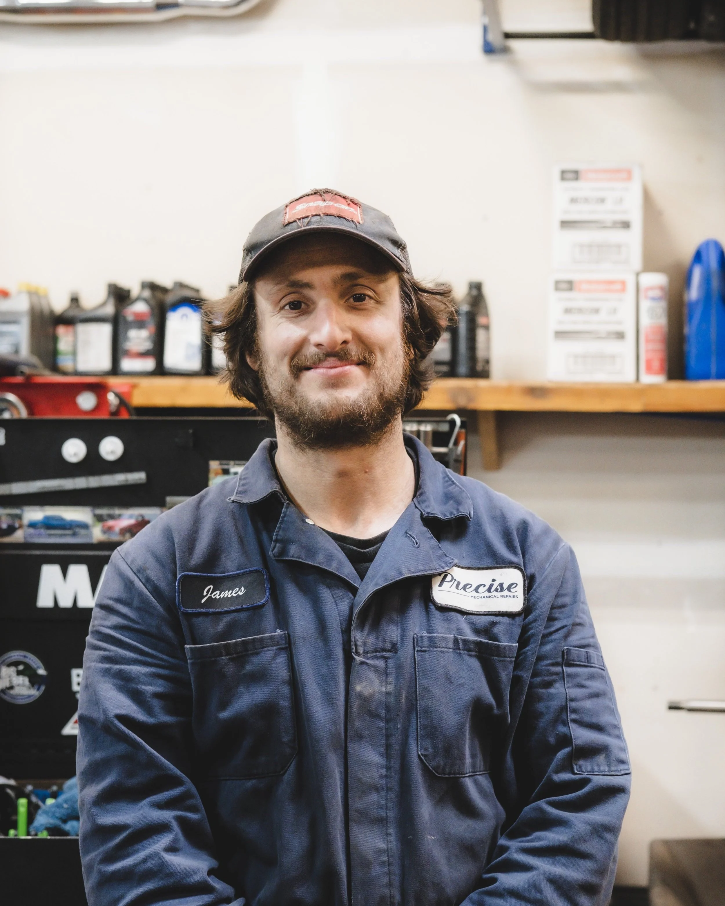 A man with a beard and wavy hair, wearing a navy blue mechanics jumpsuit with a name tag reading 'James' and a patch that says 'Precise Mechanical Repairs', standing in a garage or workshop.