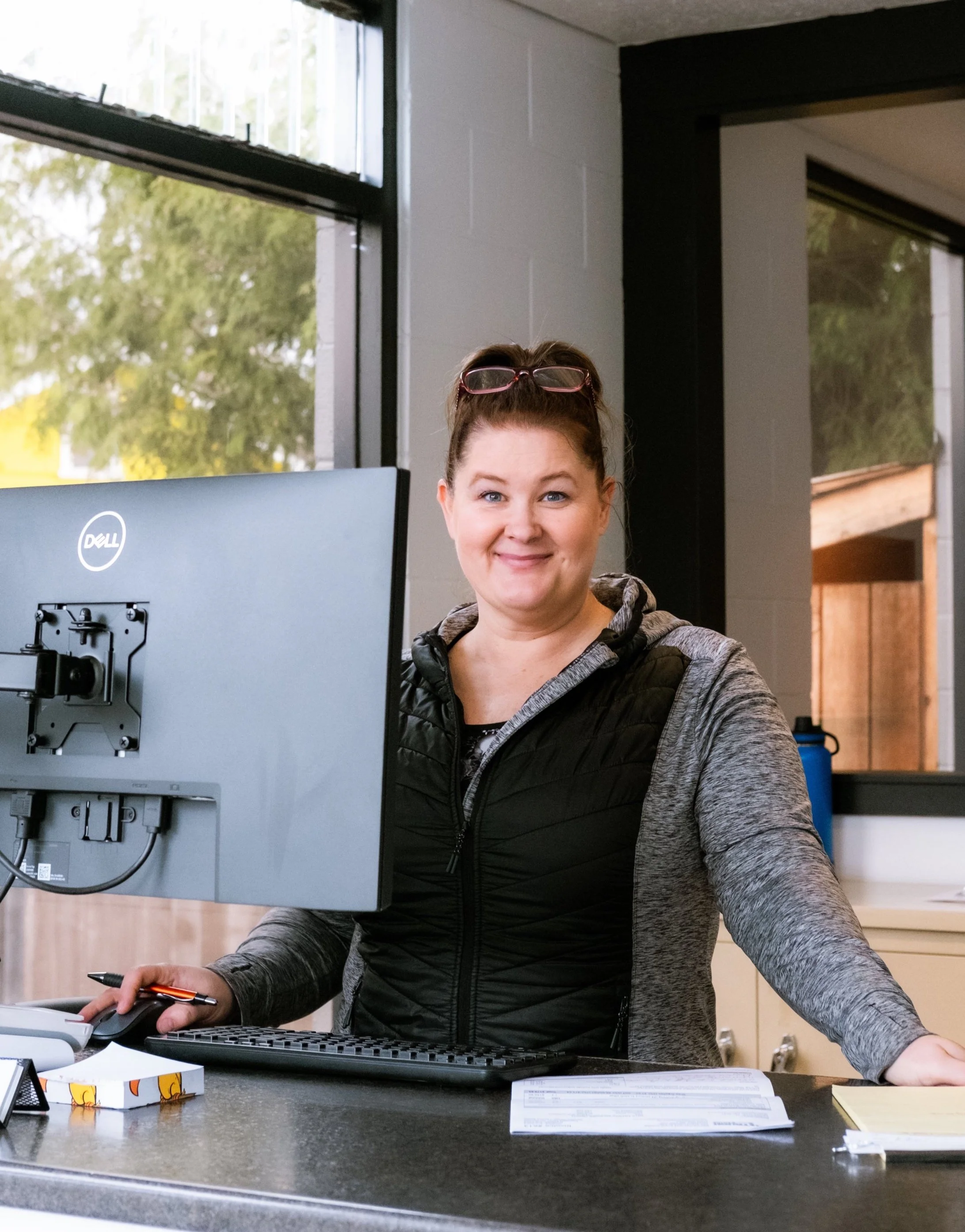 A woman smiling at a desk with a computer monitor, mouse, keyboard, and papers in an office or reception area.