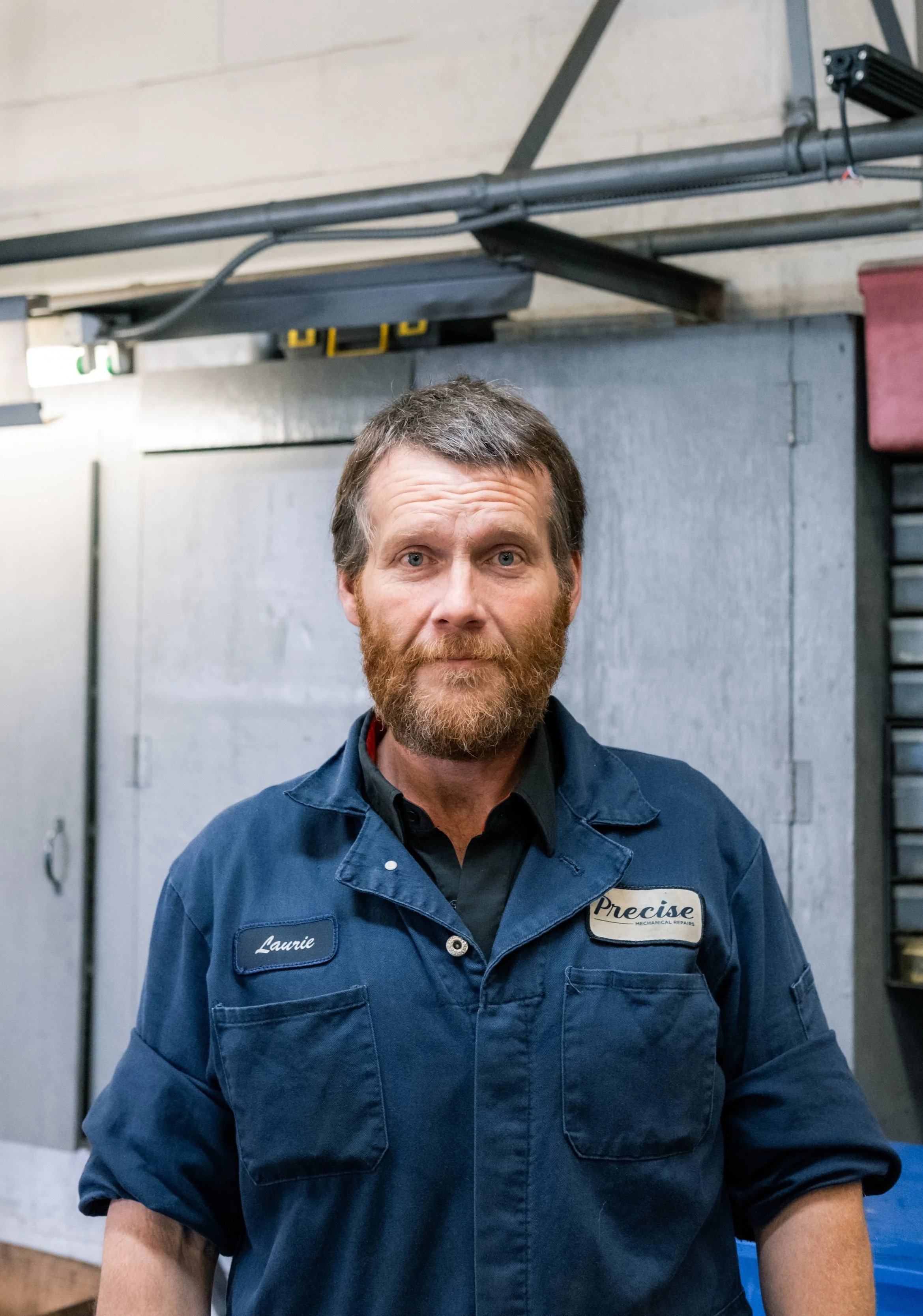 A man with a beard, wearing a blue work uniform with the name tag 'Laurie' and a patch that reads 'Precise Mechanics'