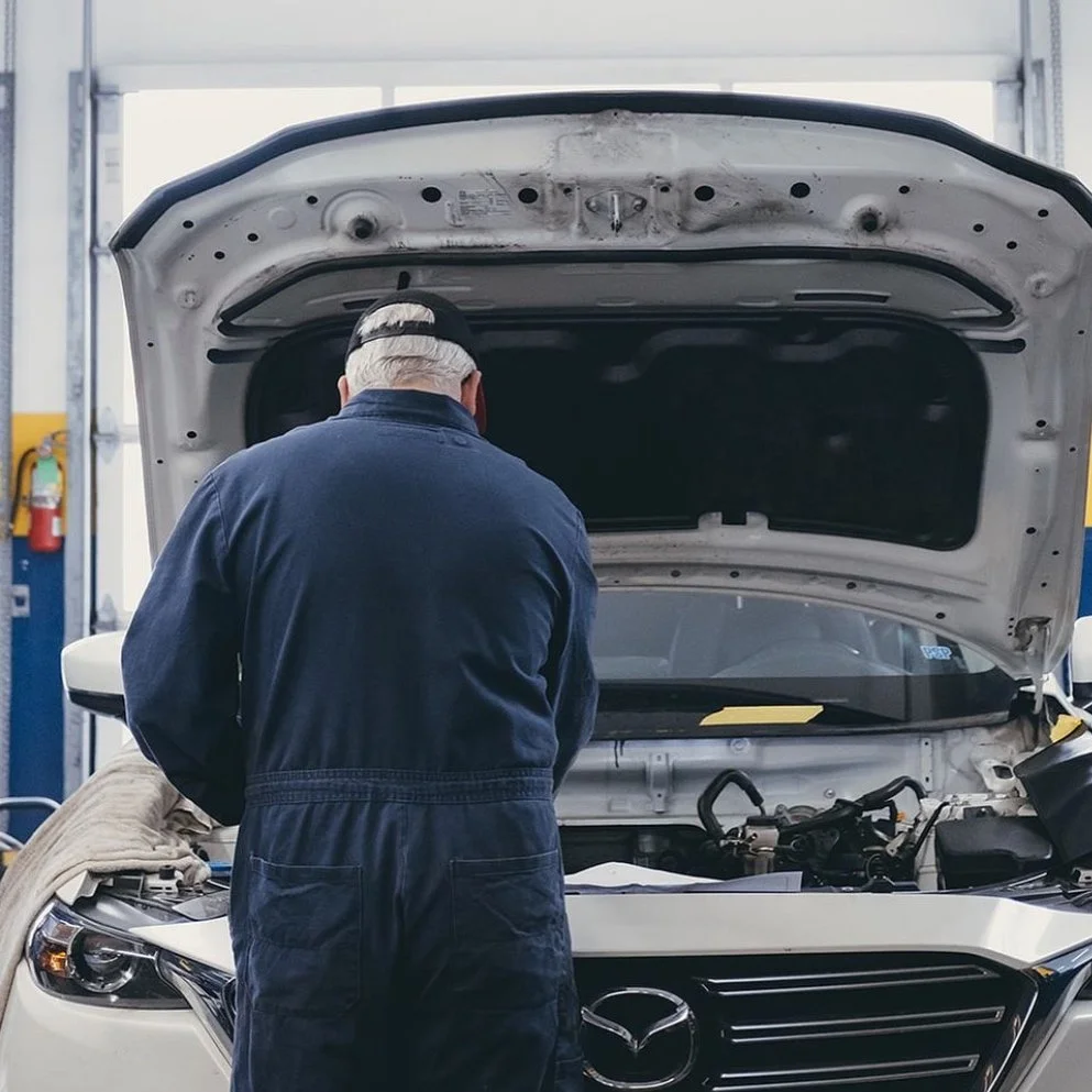 A mechanic working on a white Mazda car with the hood open in an auto repair shop.