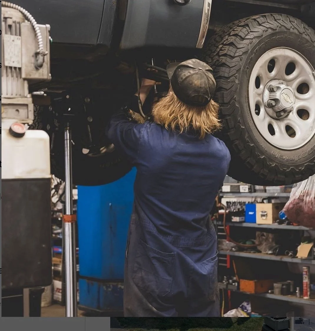 A mechanic working underneath a lifted vehicle in a garage, with the person seen from the back wearing a dark cap and coveralls.