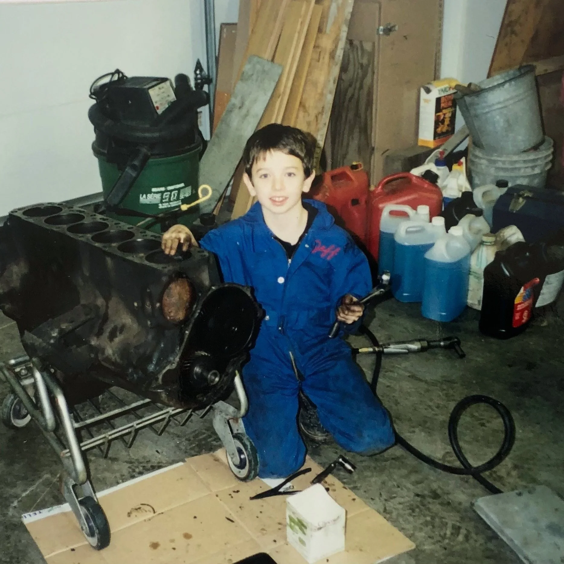 A young Jeff in a blue jumpsuit is kneeling beside a disassembled engine in a garage, holding a wrench. The garage has various tools, cans, and equipment in the background.