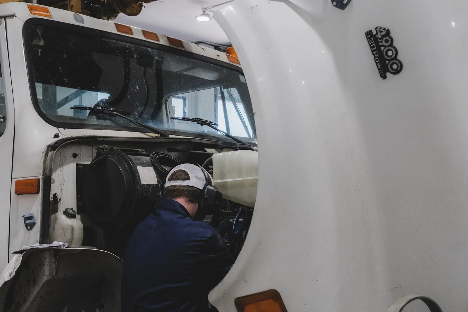 A mechanic working inside the engine compartment of a large white vehicle, possibly a bus or truck, wearing a cap and headphones.