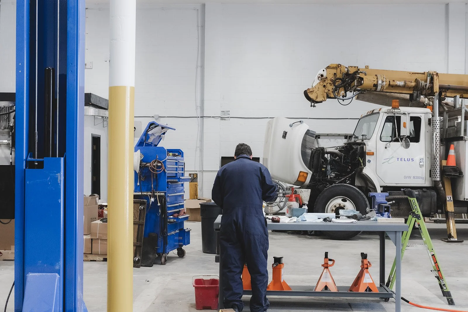 A mechanic wearing a dark jumpsuit working at a repair table in an auto repair shop, with a large white and yellow utility truck in the background.