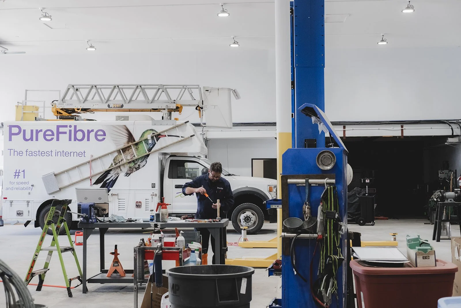 A technician working on equipment in a large industrial or service space with a vehicle inside a garage or workshop area, shelves, tools, and equipment visible.