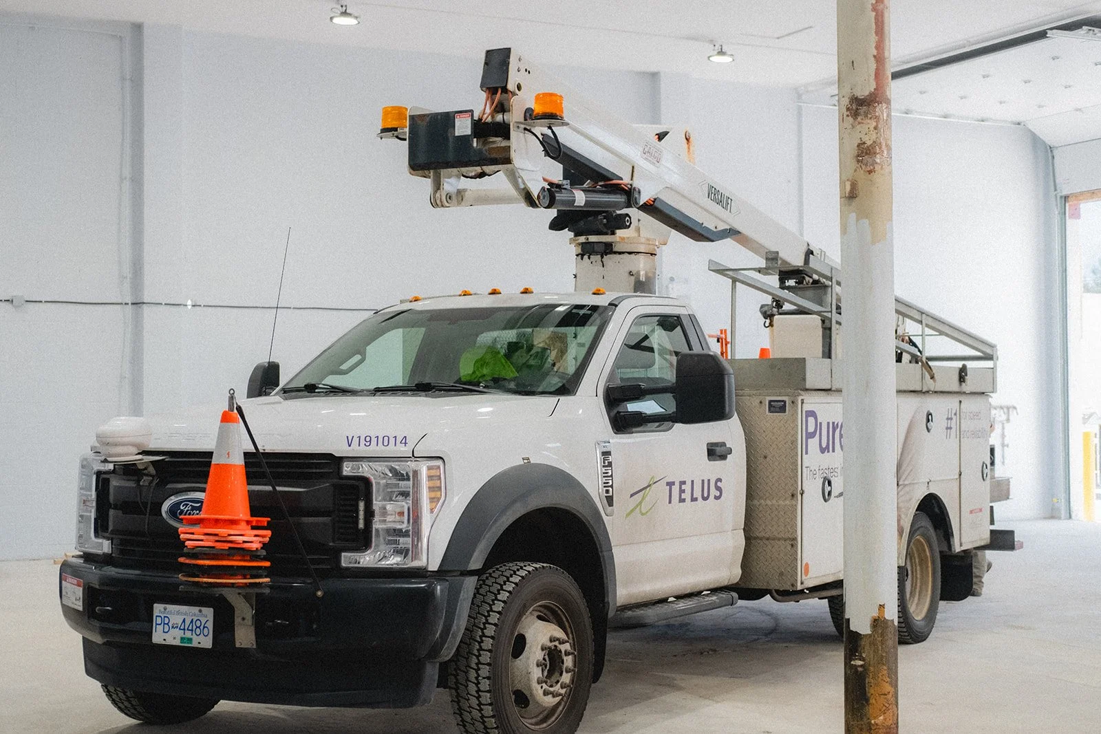 A white utility truck with TELUS branding and an orange traffic cone on the front grille, equipped with a hydraulic lift. The lift extends upwards with a platform at the end, inside a large indoor space with unfinished wall surfaces and a weathered pole.