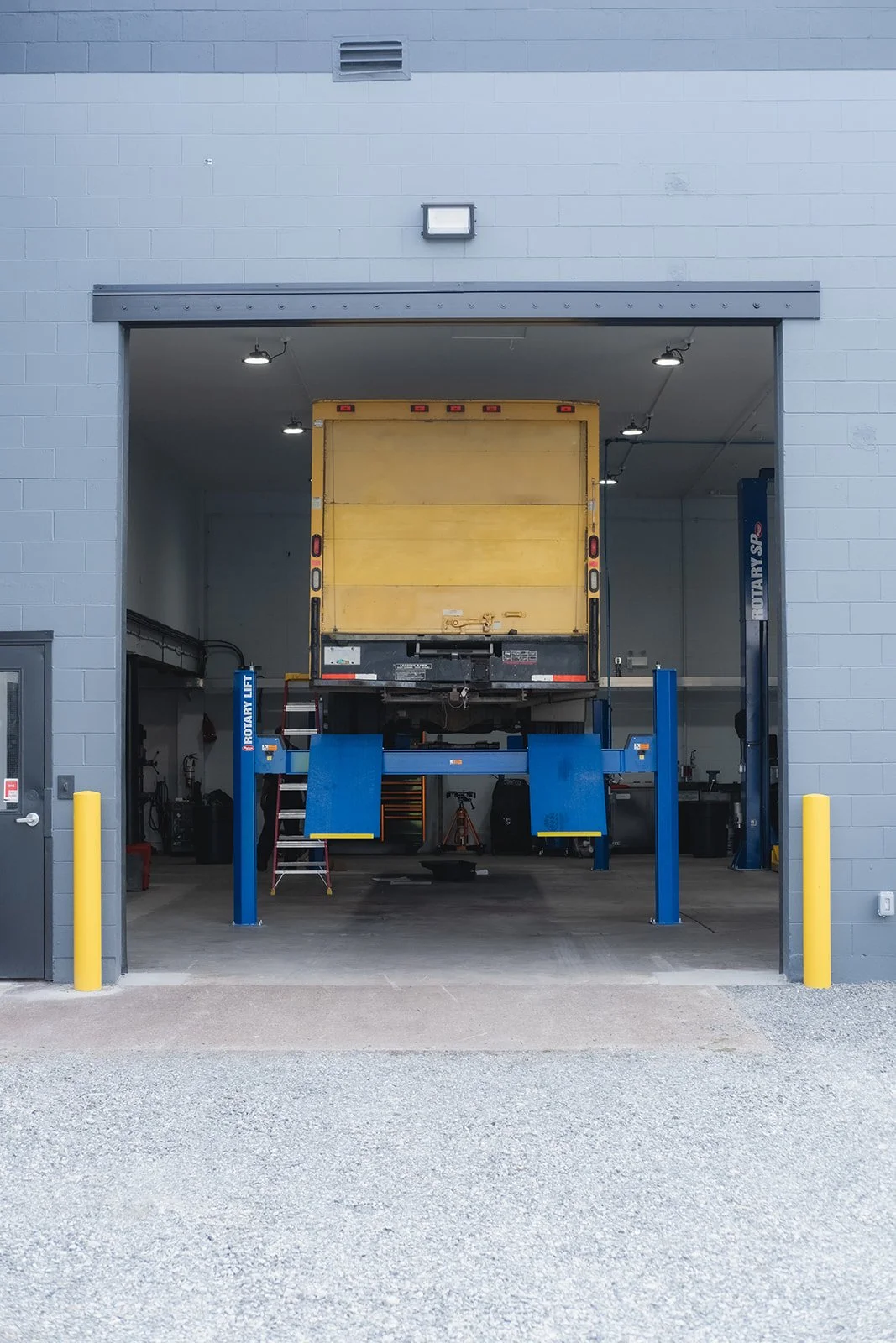 Inside an auto service garage with a yellow box truck on a blue hydraulic lift, yellow posts at the entrance, and gray walls.