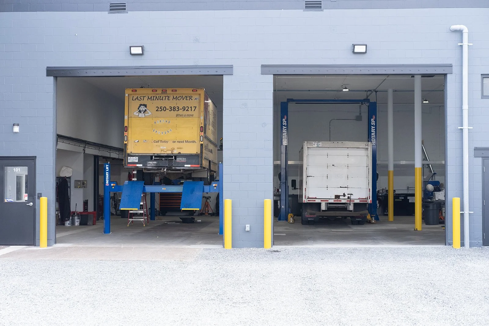 A building with two open garage bays. One bay has a yellow moving truck on a hydraulic lift. The other bay contains a white truck. The building has light gray walls, yellow bollards, and a gravel driveway in front.