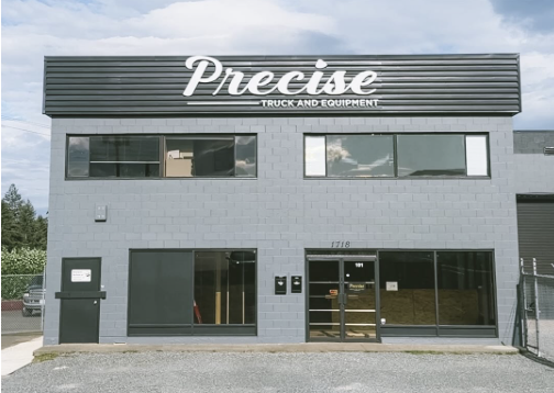 A snowy scene outside of an automotive repair shop named 'Precise Mechanical Repairs'. The shop's sign lists services including full service, transmission repairs, safety inspections, and maintenance. Several cars are parked in front, with a background of snow-covered trees and a building tower.