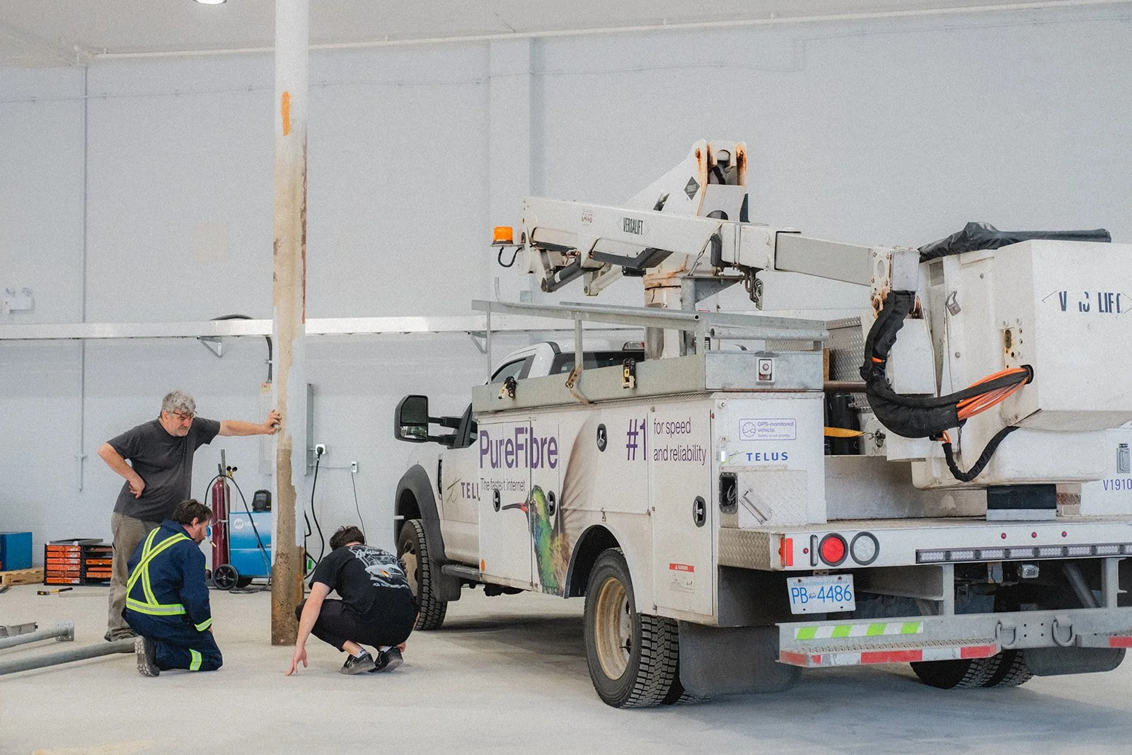 Three workers inspecting a utility truck with an extended cherry picker in a warehouse or garage. One worker is leaning on a pole, another is squatting, and the third is standing.